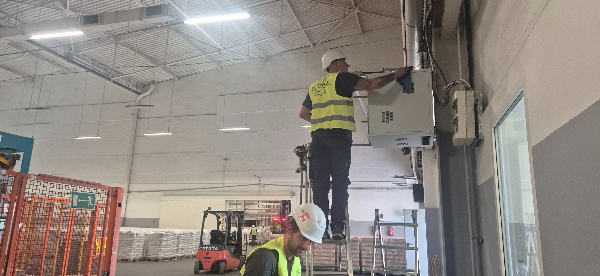 Pracownik w kasku i kamizelce na drabinie czyści szafę elektryczną w hali magazynowej. Widoczne kable, skrzynki i oświetlenie przemysłowe.
