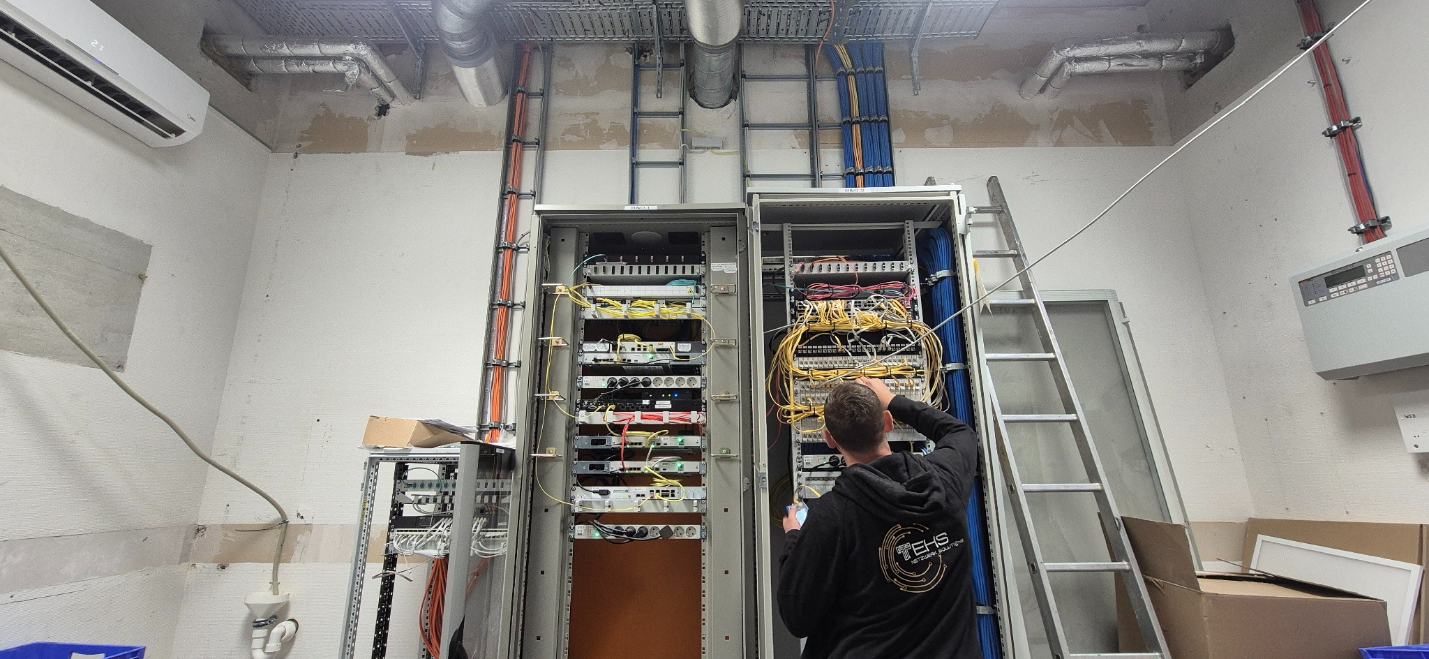 Pracownik w czarnej bluzie konfiguruje kable w szafie rack z urządzeniami sieciowymi. Widoczne kable, przełączniki, drabina i instalacje pod sufitem.