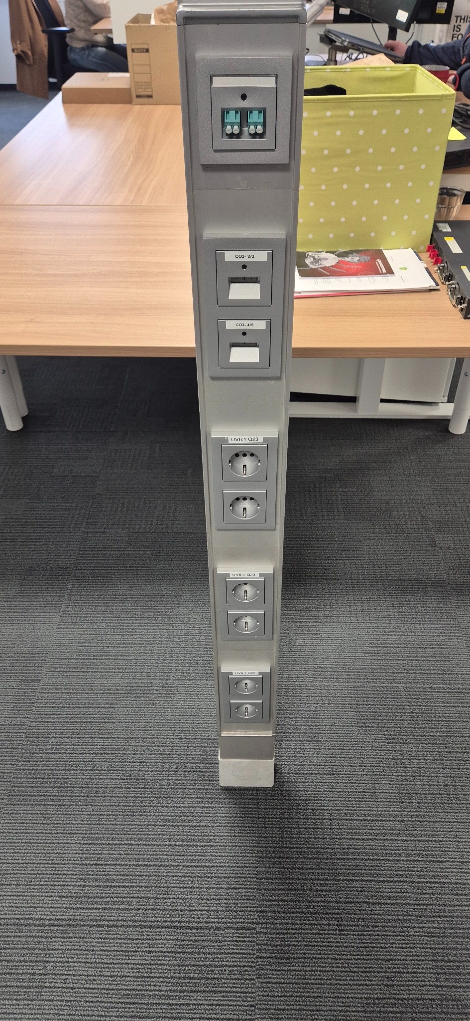 Srebrny słup z gniazdkami elektrycznymi i portami sieciowymi w biurze, widok z bliska na szarym dywanie. Gniazdka oznaczone etykietami.