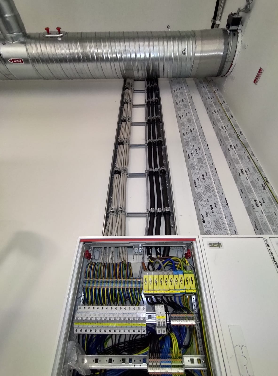 Rozdzielnia elektryczna z okablowaniem ułożonym w korytach na ścianie. Widoczne przewody w różnych kolorach i grubościach, oraz elementy zabezpieczające.