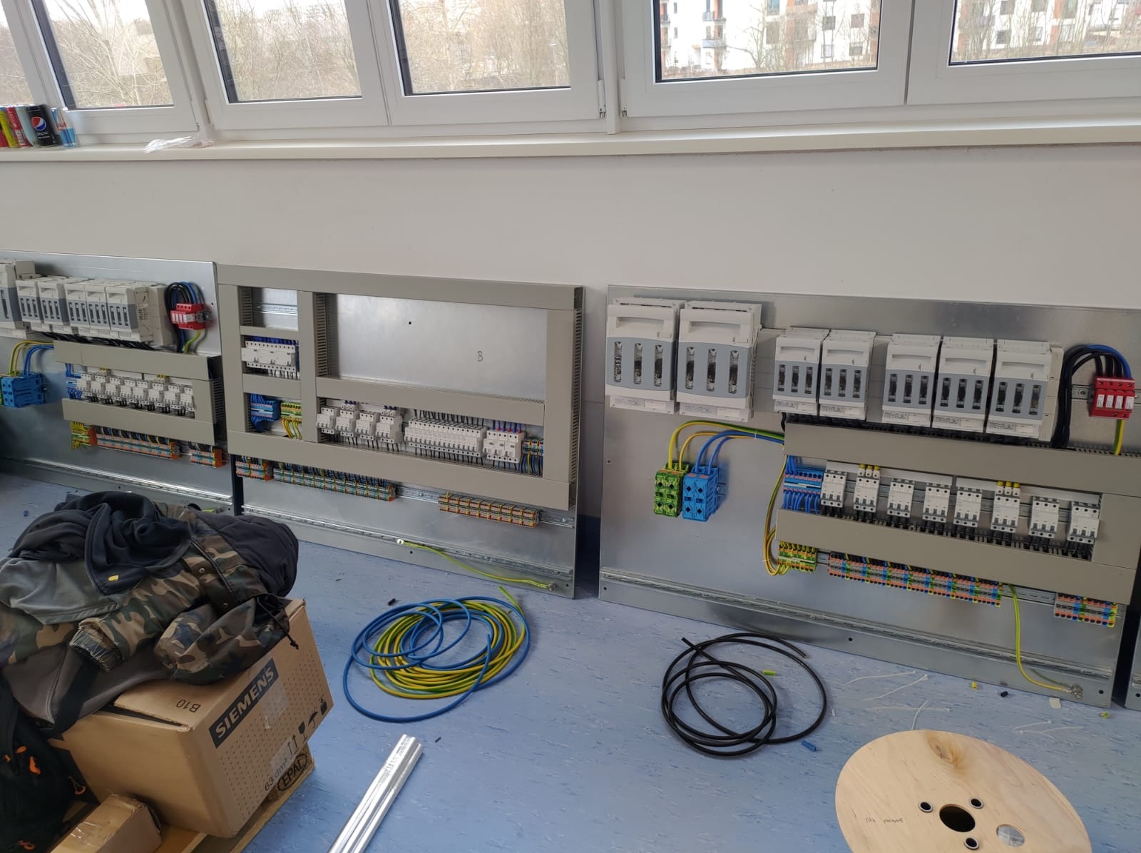Rozdzielnia elektryczna w trakcie montażu, widoczne kable, bezpieczniki i elementy sterujące. Na podłodze leżą zwoje kabli i pusta szpula.
