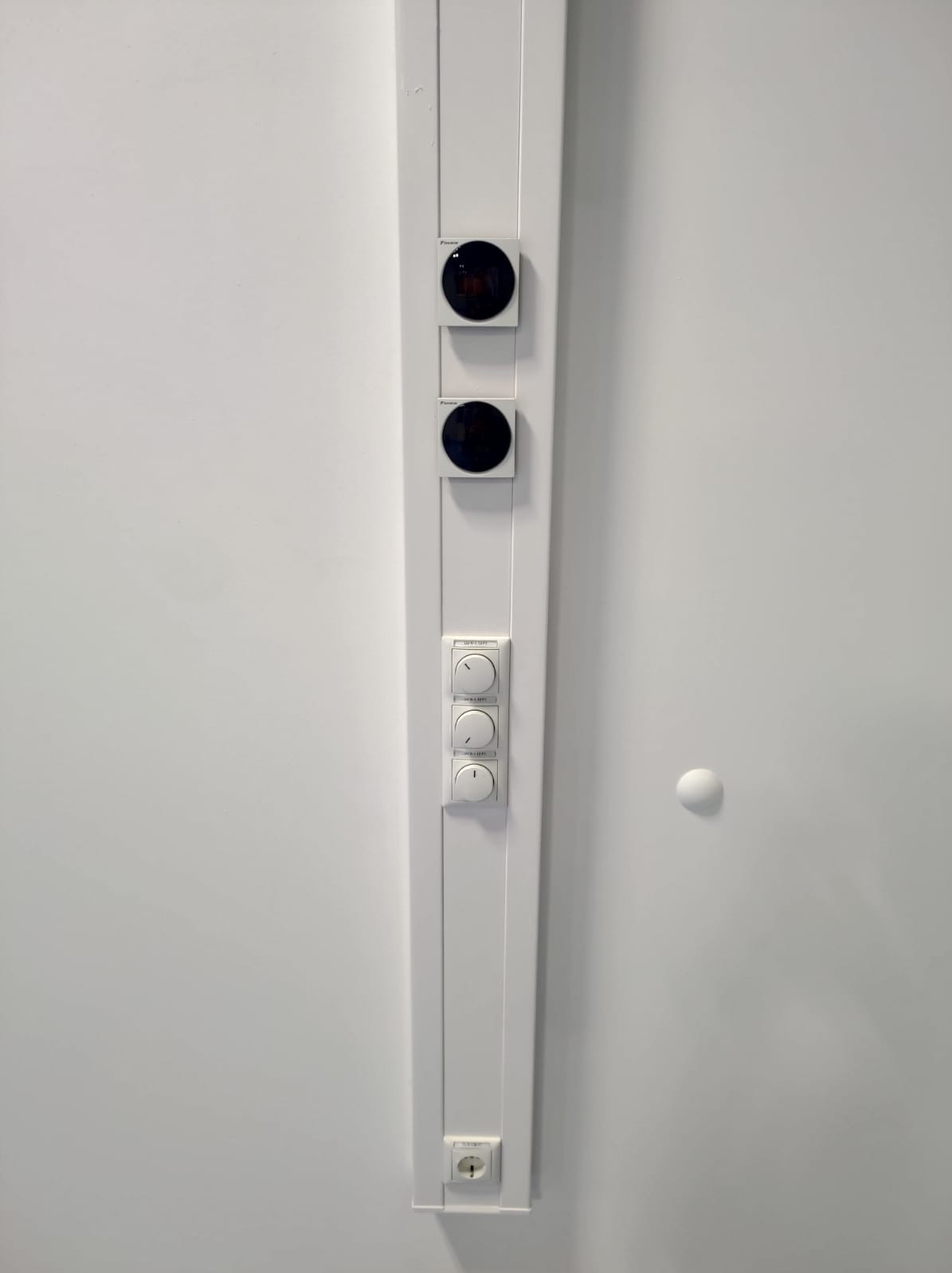 Biała listwa z włącznikami światła, gniazdkiem i dwoma okrągłymi czujnikami na białej ścianie. Minimalistyczny design, nowoczesne wykończenie.