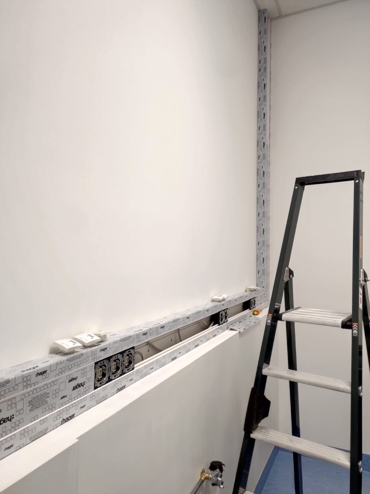 Montaż podtynkowych gniazdek elektrycznych w listwie przypodłogowej, widoczna drabina i fragment instalacji w rogu pomieszczenia.