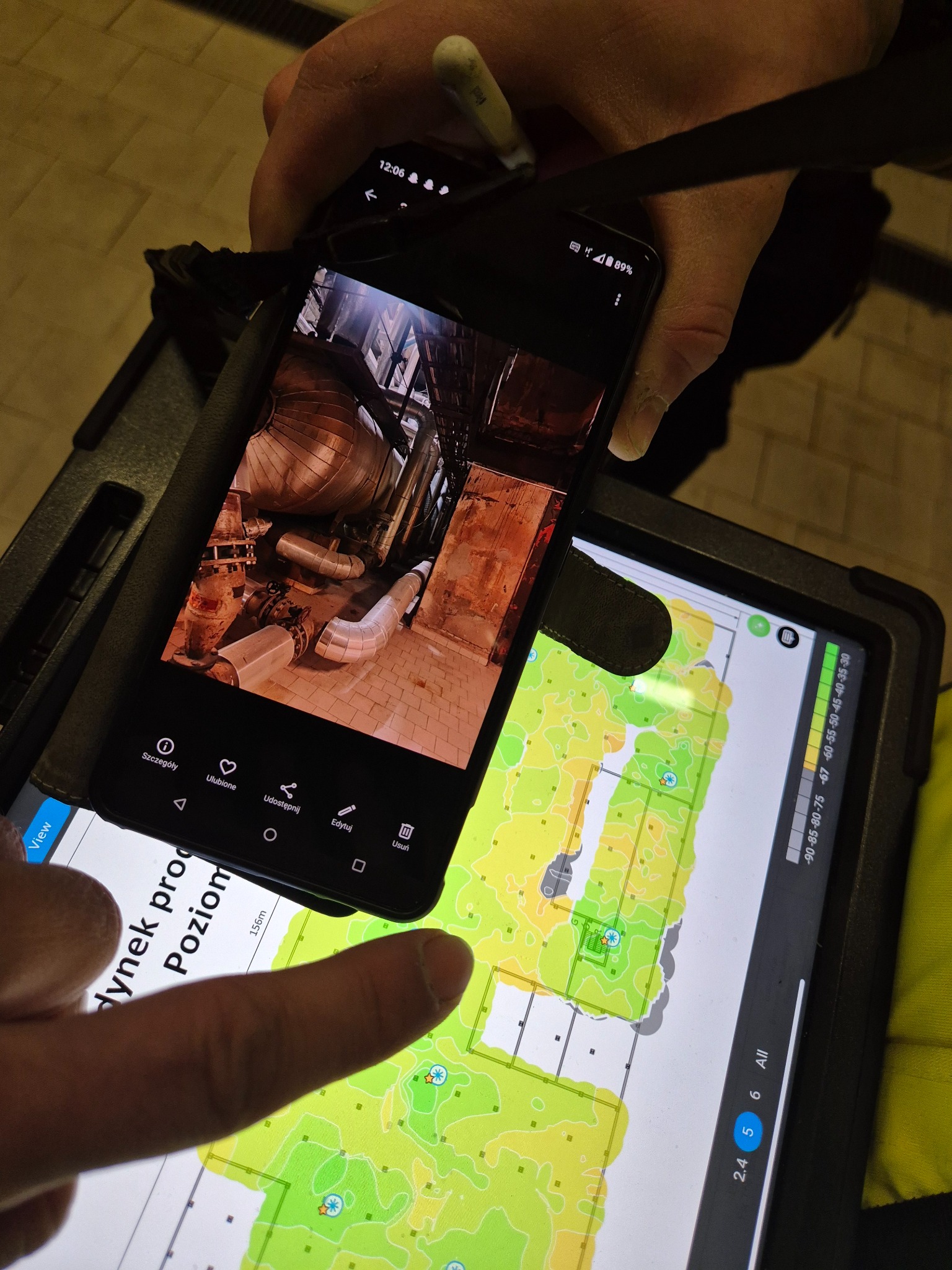 Zdjęcie rurociągu przemysłowego na ekranie smartfona, trzymanego nad tabletem z planem budynku, wskazywanym palcem. Analiza danych na budowie.