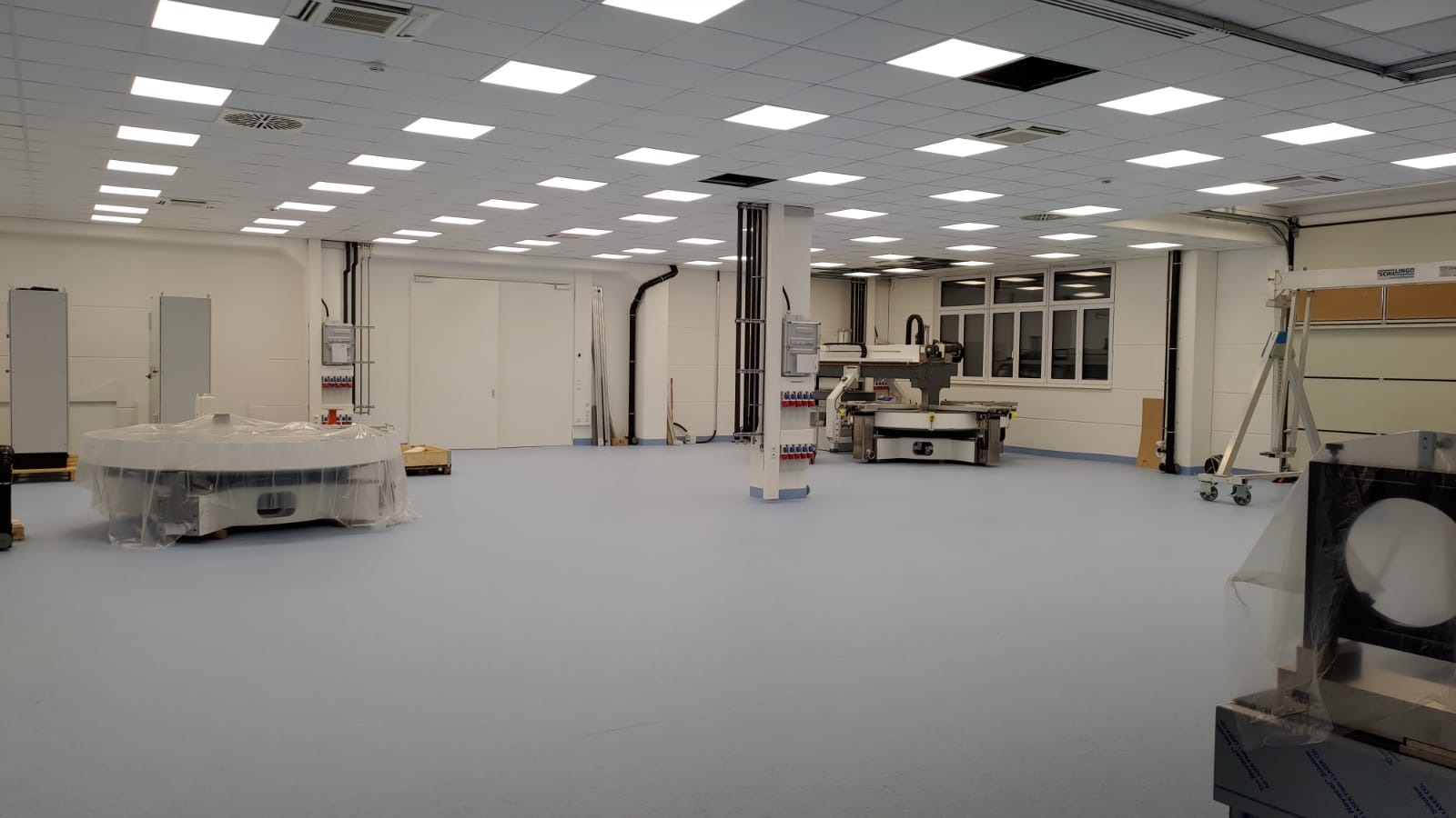 Jasne, przestronne pomieszczenie z równomiernym oświetleniem sufitowym, widoczne urządzenia i instalacje elektryczne na ścianach. Minimalistyczny wystrój, dominują biel i błękit.