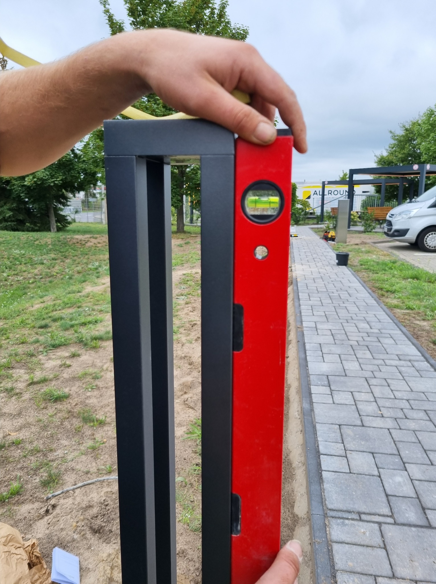 Poziomowanie słupka ogrodzeniowego z użyciem poziomicy w kolorze czerwonym. Widoczny fragment chodnika z kostki brukowej i trawnik w tle.