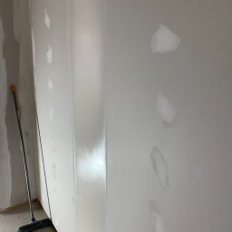 OLEH PYLYPCHAK - Ściana po szpachlowaniu z widocznymi łatami i śladami po szpachli. W rogu stoi miotła, co sugeruje prace porządkowe po zakończonym etapie prac.