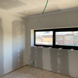 OLEH PYLYPCHAK - Pokój w trakcie remontu: ściany z częściowo nałożoną gładzią, okno z czarną ramą, widoczne przewody elektryczne. Podłoga wyłożona panelami. Etap przygotowania do malowania.