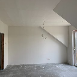 OLEH PYLYPCHAK - Poddasze po gładziowaniu ścian i sufitu, widoczne kable elektryczne, surowa ościeżnica drzwi i okno balkonowe z widokiem na zewnątrz. Szary beton na podłodze.