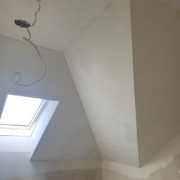 OLEH PYLYPCHAK - Poddasze po gładziowaniu z oknem dachowym i wystającym kablem oświetleniowym. Ściany i sufit w jasnym odcieniu bieli, gotowe do malowania.