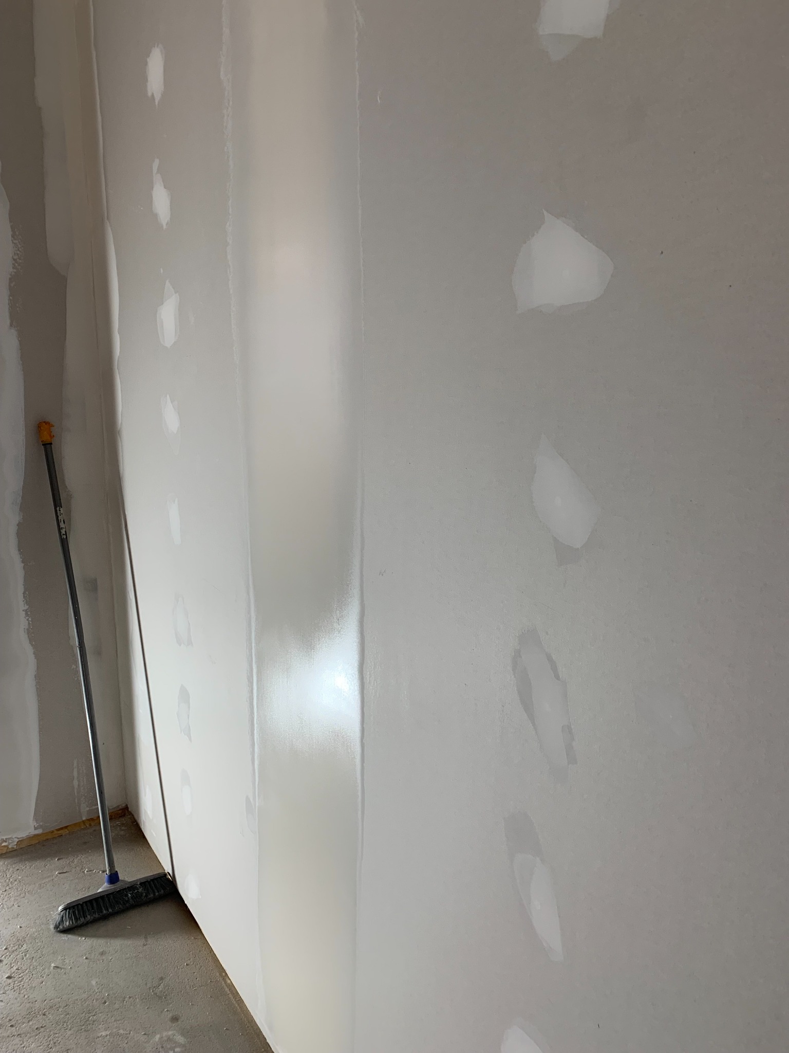 Ściana po szpachlowaniu z widocznymi łatami i śladami po szpachli. W rogu stoi miotła, co sugeruje prace porządkowe po zakończonym etapie prac.