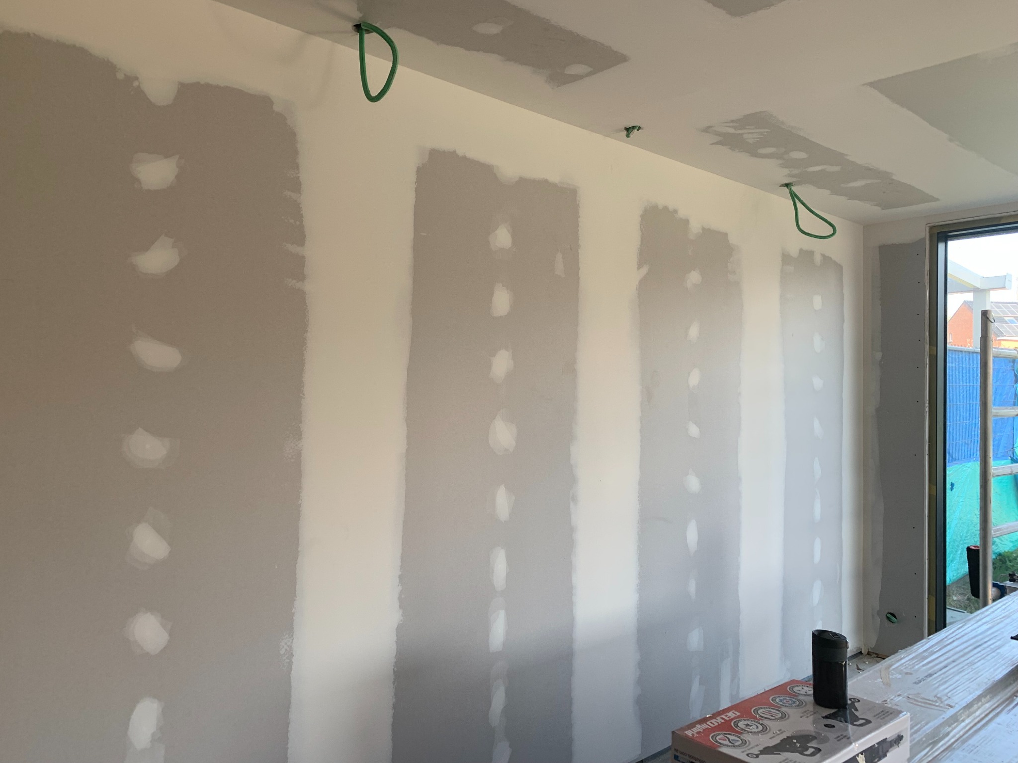 Ściana z płyt kartonowo-gipsowych w trakcie szpachlowania, widoczne łączenia i otwory po wkrętach. Na podłodze leżą płyty i narzędzia.