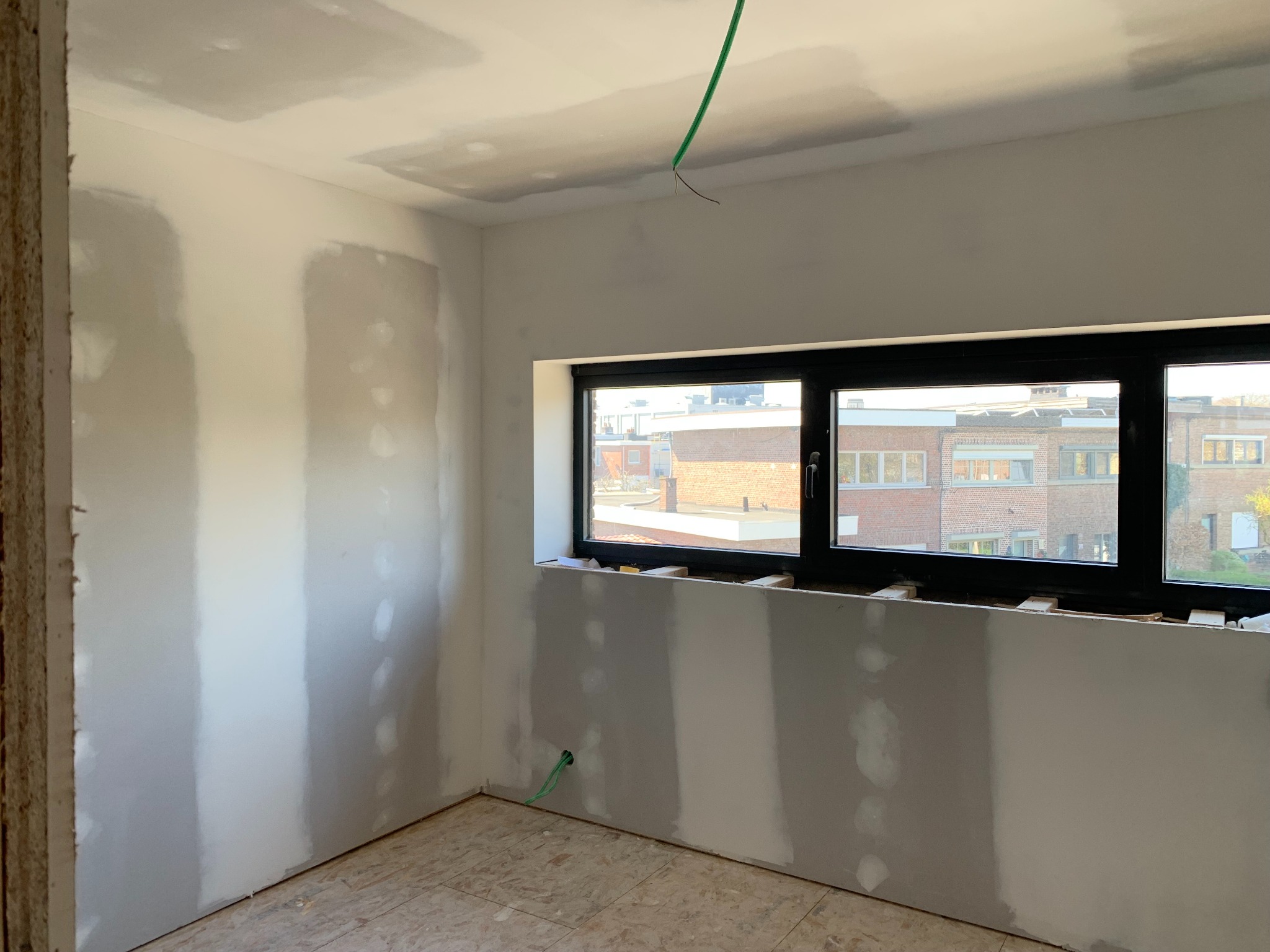 Pokój w trakcie remontu: ściany z częściowo nałożoną gładzią, okno z czarną ramą, widoczne przewody elektryczne. Podłoga wyłożona panelami. Etap przygotowania do malowania.
