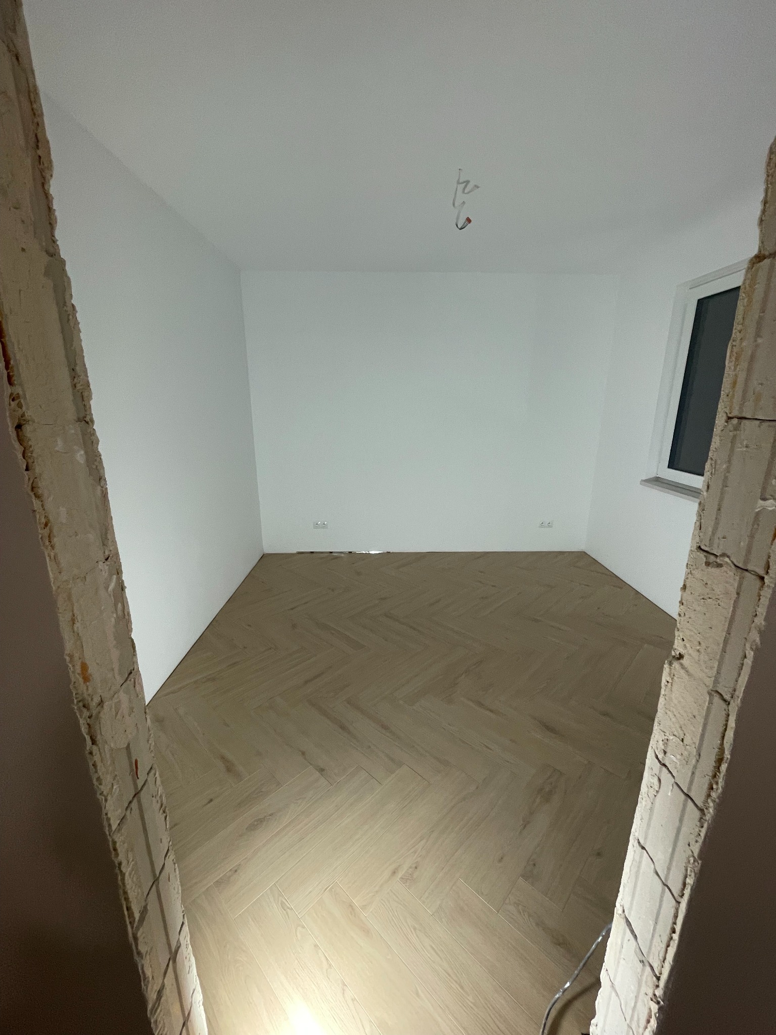 Pokój po remoncie z parkietem w jodełkę, widok przez surowy otwór drzwiowy. Świeżo pomalowane białe ściany, okno z parapetem. Wystające kable na suficie.