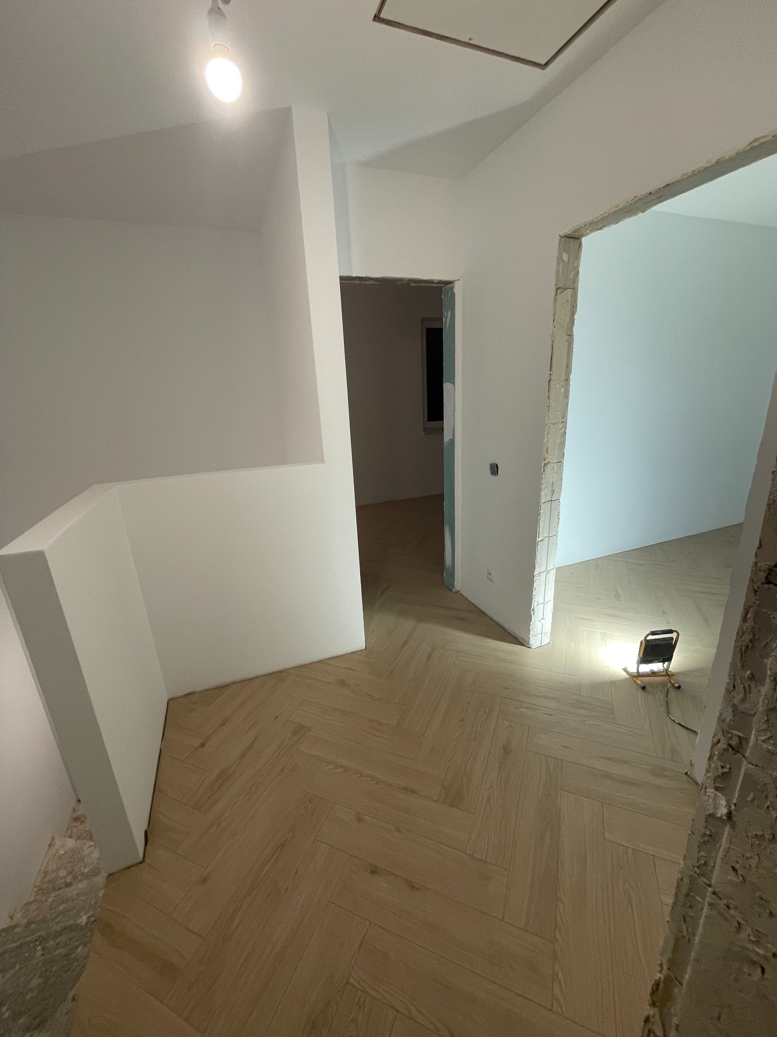 Jasne, drewnopodobne panele ułożone w jodełkę na podłodze w remontowanym wnętrzu. Widoczne surowe ściany i ościeżnice, lampa budowlana oświetla przestrzeń.