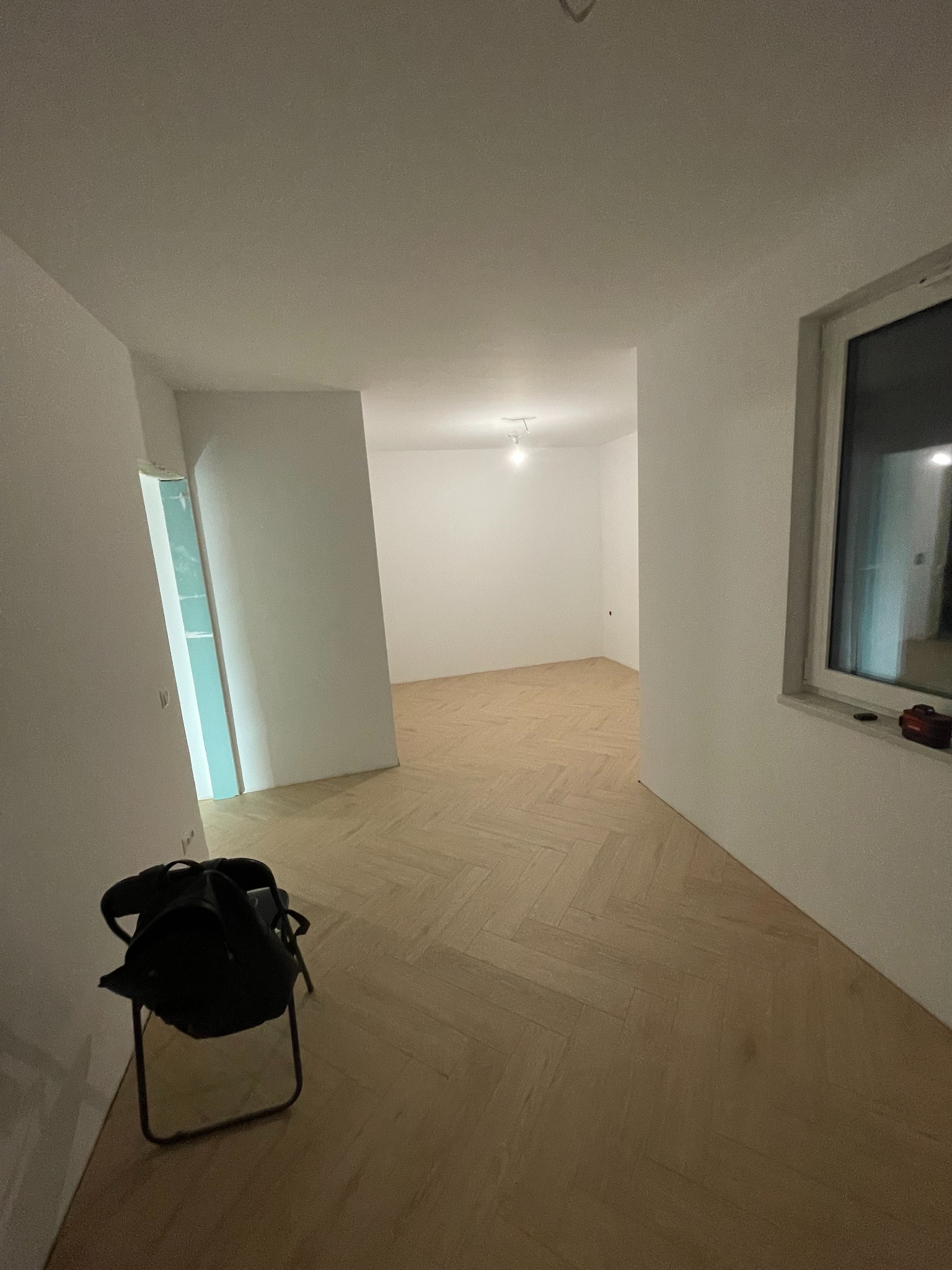 Jasne, wykończone wnętrze z parkietem w jodełkę. Widoczne białe ściany, okno i krzesło z torbą. W głębi pomieszczenia widoczne oświetlenie sufitowe.