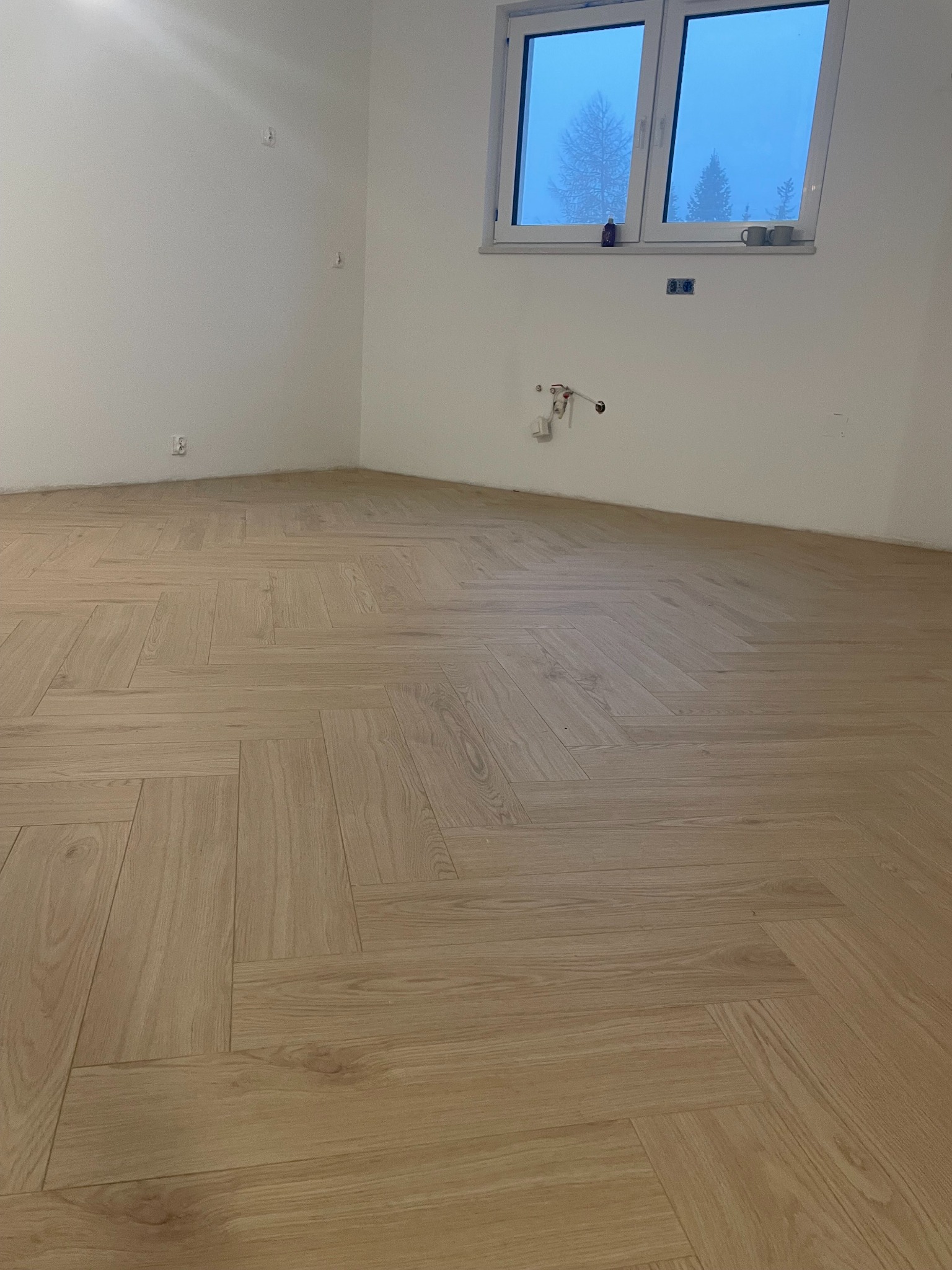 Ukończona podłoga z parkietu ułożonego w jodełkę w jasnym, pustym pokoju z oknem. Widoczne przygotowane instalacje na ścianie.