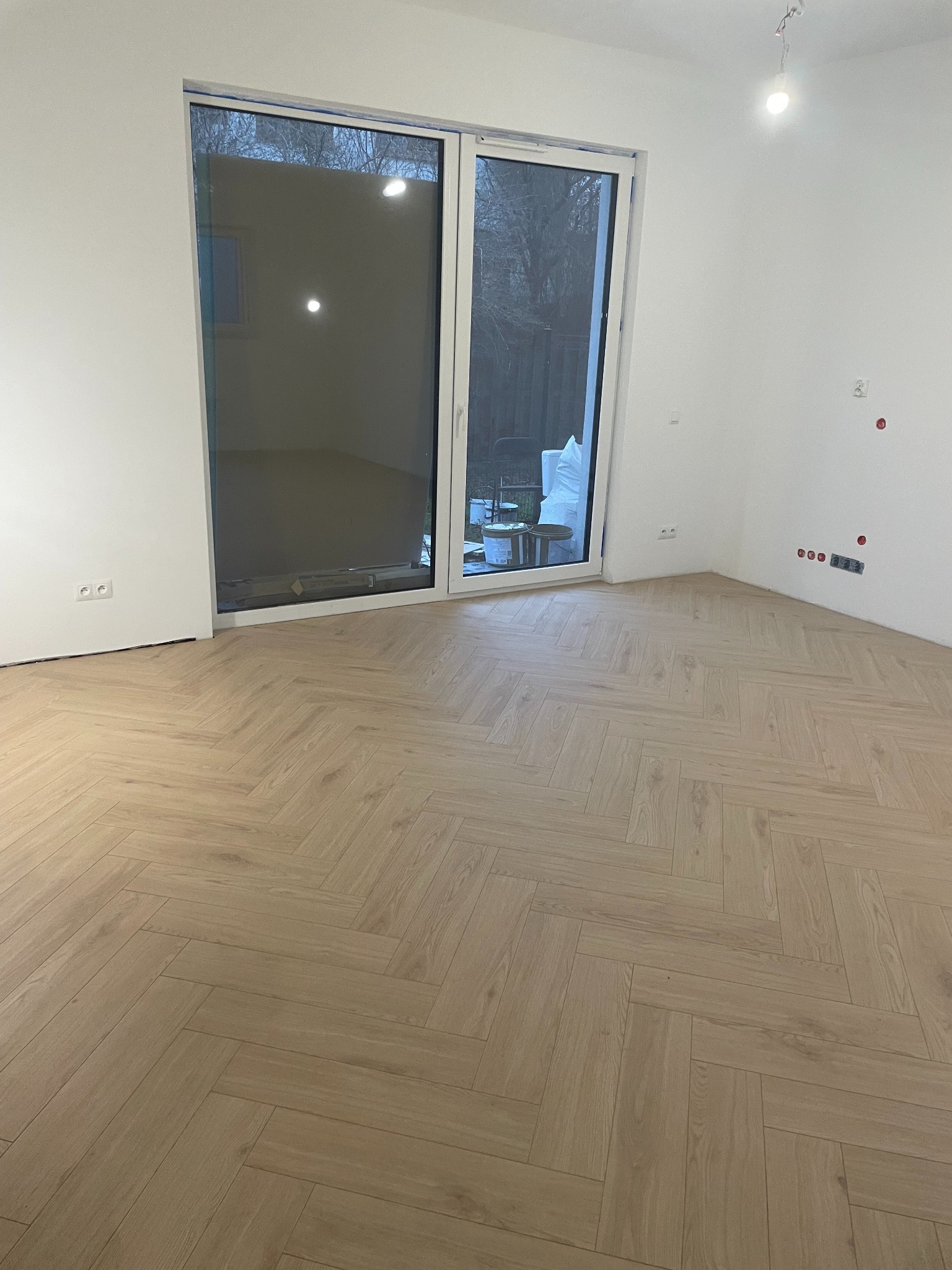 Jasna podłoga ułożona w jodełkę w nowym pokoju z dużym oknem balkonowym. Widoczne puszki elektryczne i gniazdka. Świeżo wykończone ściany.