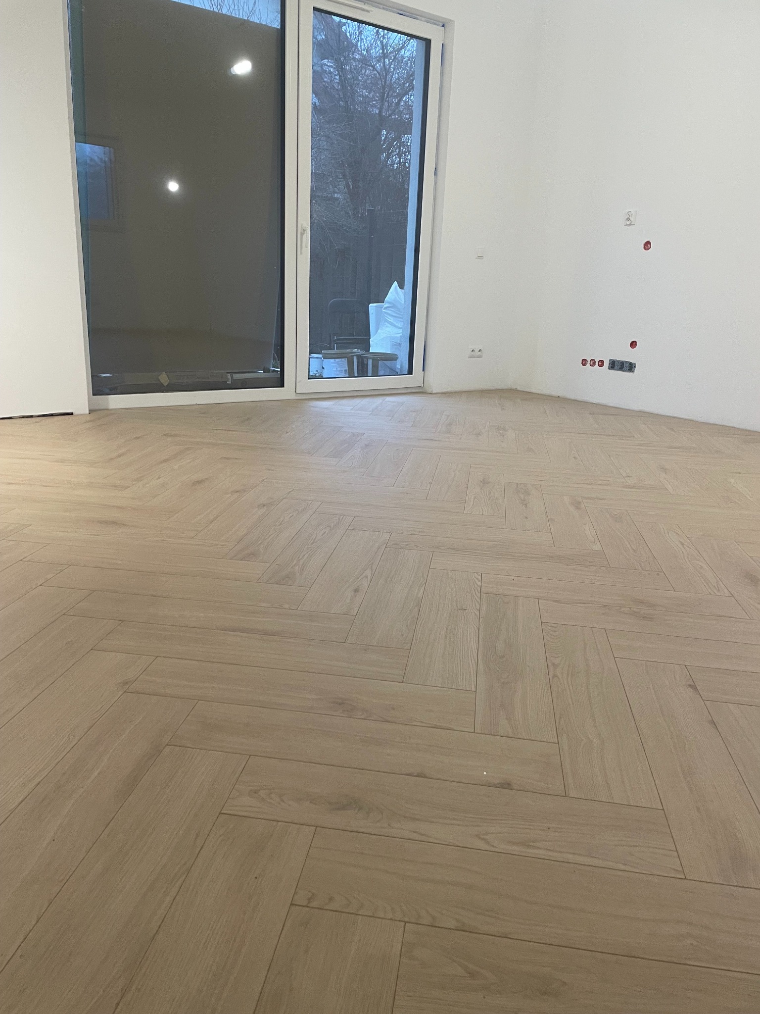Jasna podłoga ułożona w jodełkę w nowym wnętrzu z dużym oknem balkonowym i przygotowanymi punktami elektrycznymi na ścianie.