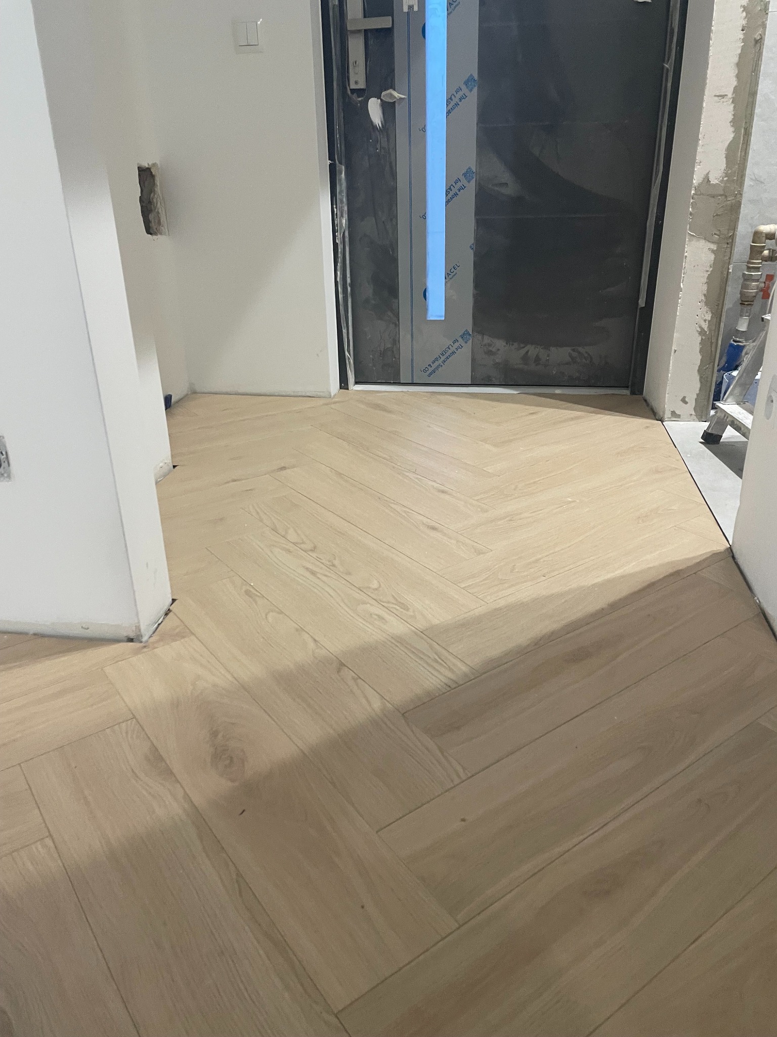 Ułożona podłoga z paneli w jodełkę, widok z góry. Drzwi wejściowe z folią ochronną i fragment ściany z niedokończonym gniazdkiem elektrycznym w tle.