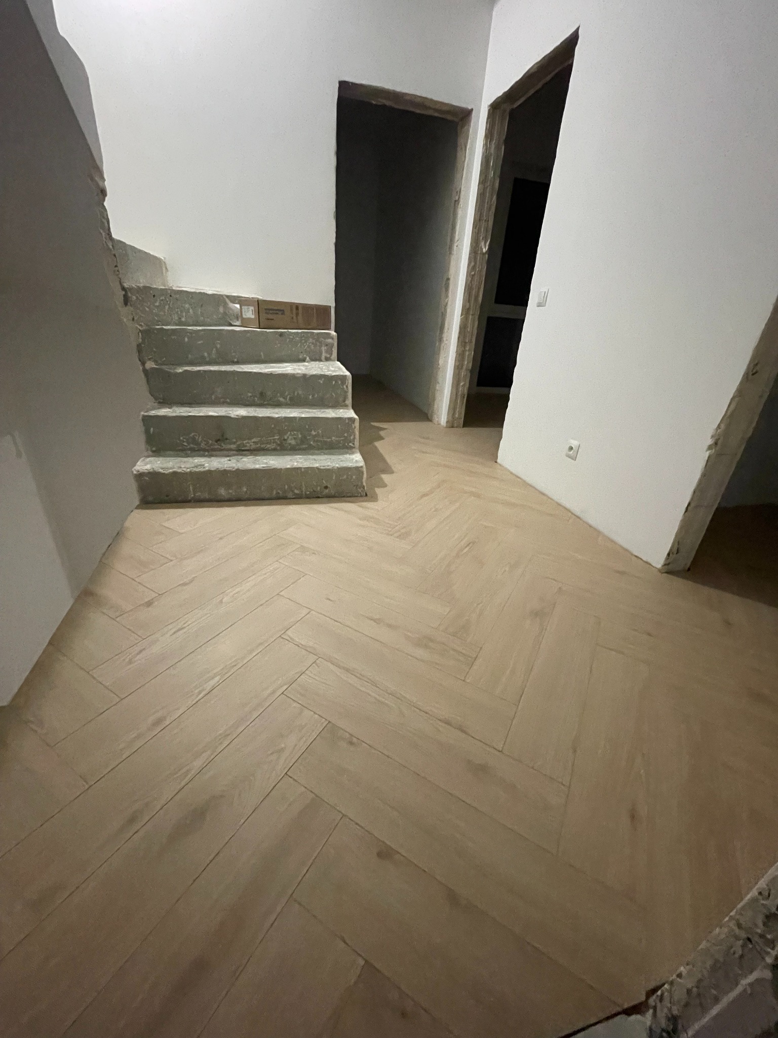 Ułożona podłoga z paneli w jodełkę, surowe schody betonowe, widoczne przygotowania do wykończenia ścian i futryn drzwiowych. Wnętrze w trakcie remontu.