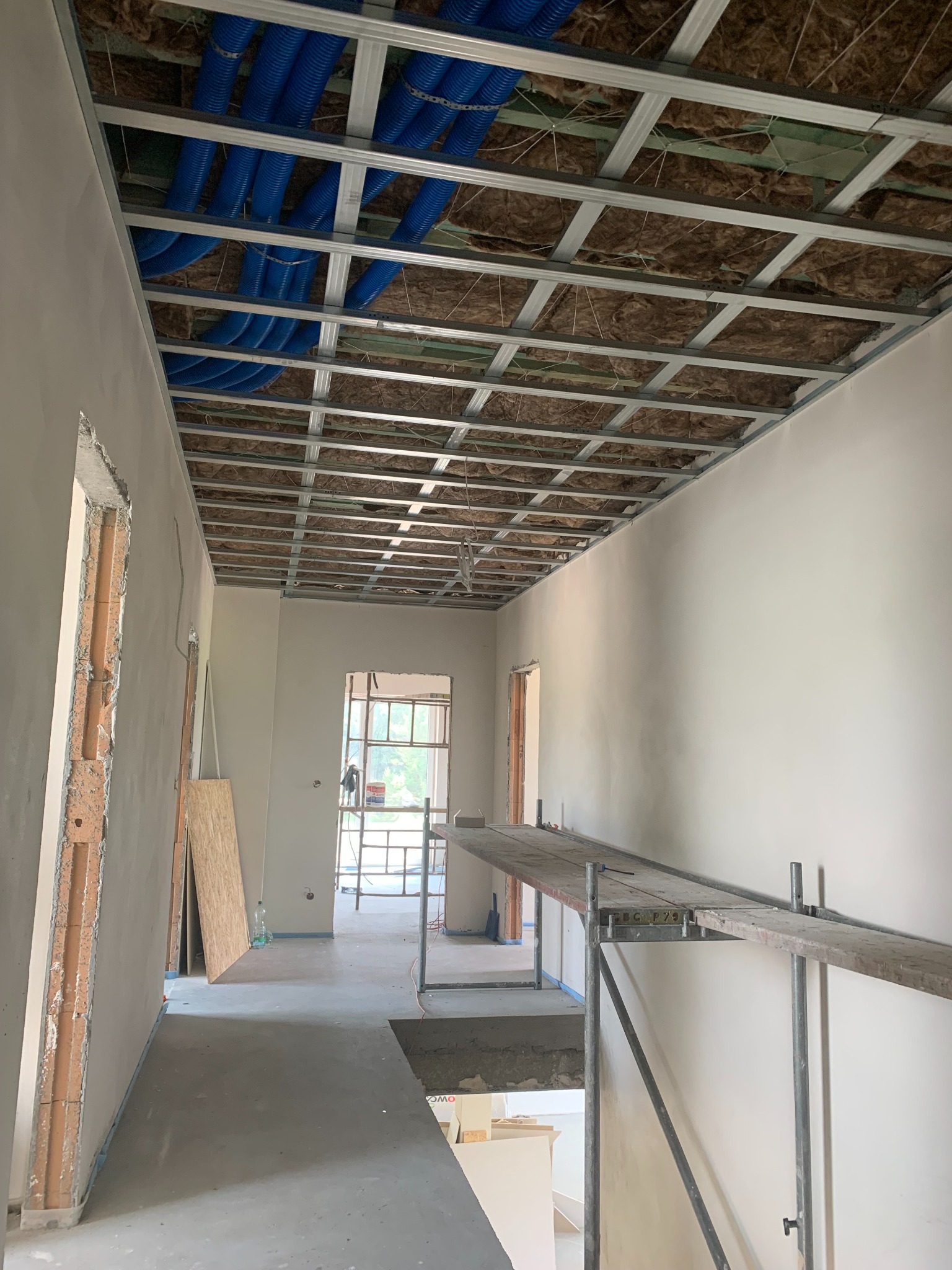 Wnętrze w trakcie prac wykończeniowych: sufit podwieszany z izolacją, instalacja wentylacyjna, ściany przygotowane do malowania, rusztowanie. Widok na korytarz.