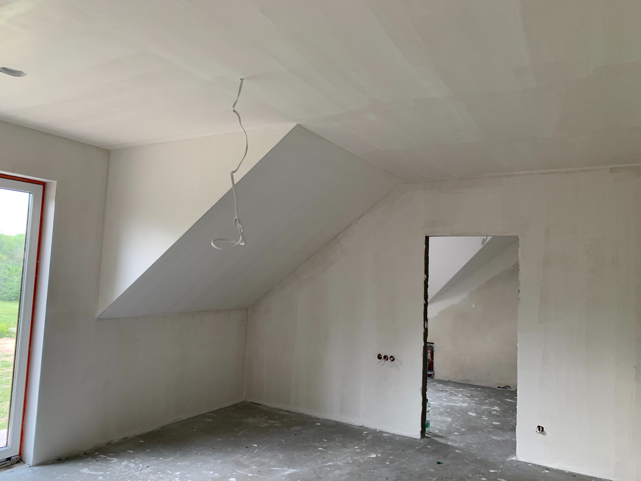 Pokój na poddaszu w trakcie prac wykończeniowych: ściany i sufit pokryte gładzią, widoczne kable, okno z widokiem na zieleń, surowy beton na podłodze.