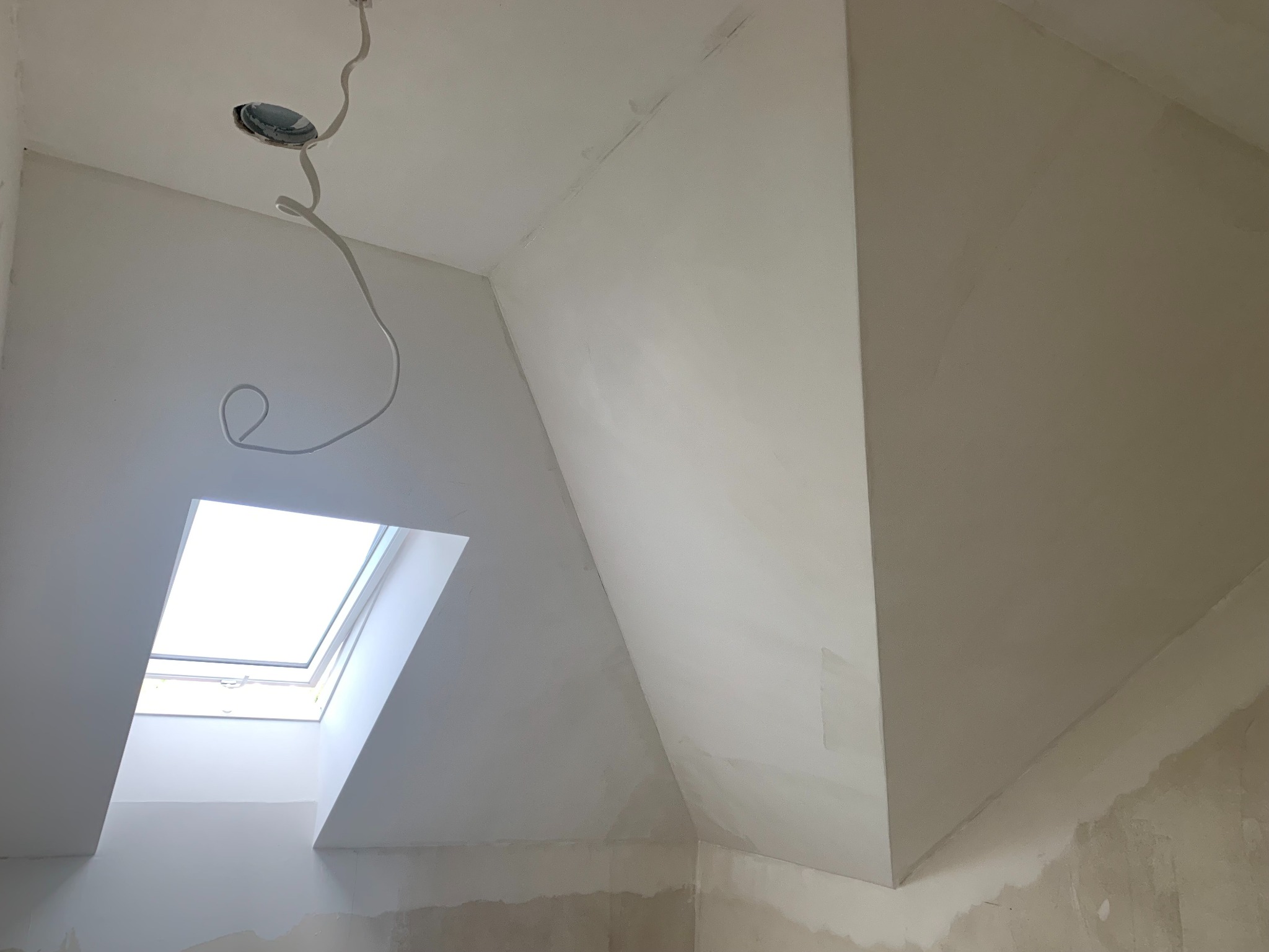 Poddasze po gładziowaniu z oknem dachowym i wystającym kablem oświetleniowym. Ściany i sufit w jasnym odcieniu bieli, gotowe do malowania.