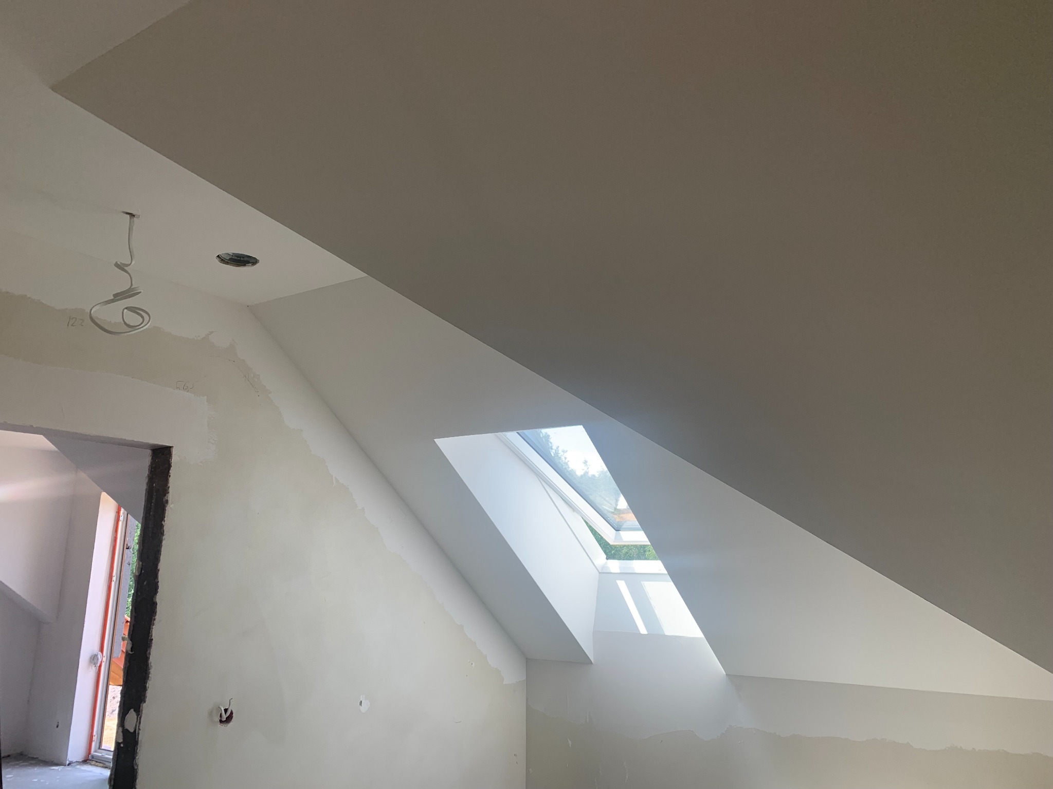 Poddasze ze świeżo wykończonym oknem dachowym, ściany przygotowane do malowania, widoczne ślady szpachlowania. Otwór drzwiowy w tle. W suficie otwór na oświetlenie.