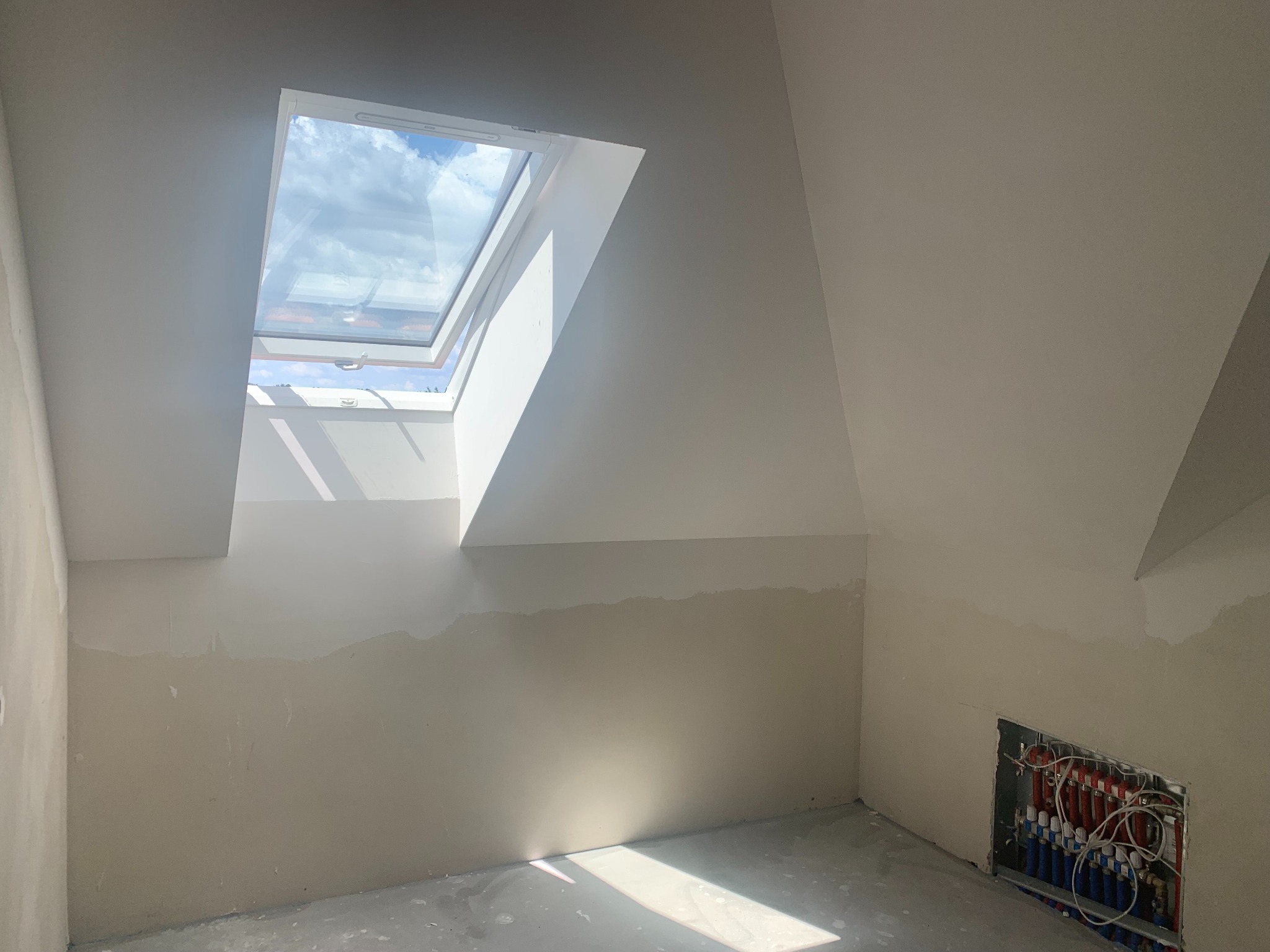 Poddasze w trakcie prac wykończeniowych: okno dachowe, ściany częściowo wyszpachlowane, instalacja grzewcza podłogowa w ścianie. Widok na niebo przez okno.