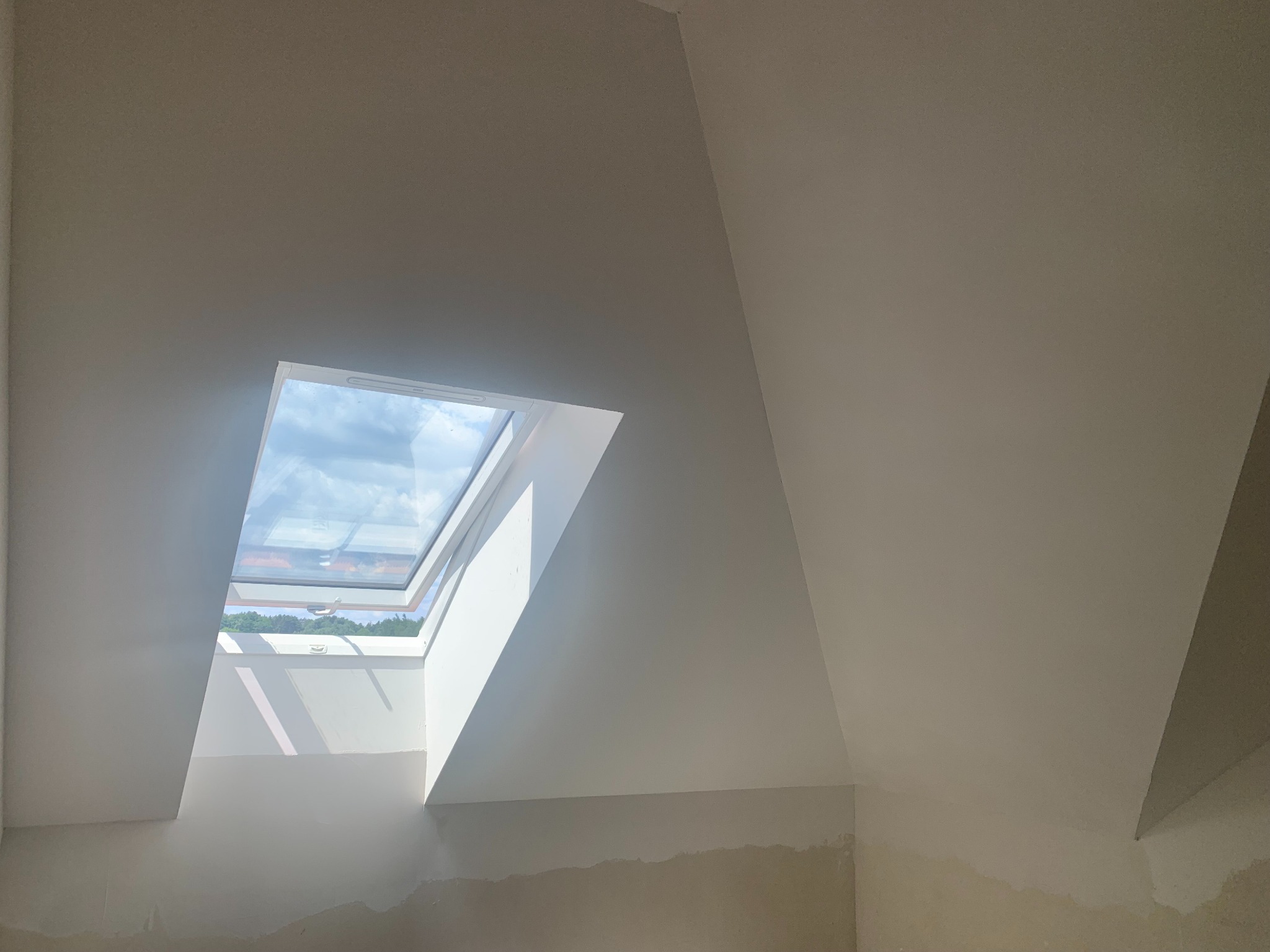 Białe, świeżo pomalowane poddasze z oknem dachowym, przez które widać błękitne niebo. Dolna część ściany przygotowana do dalszych prac wykończeniowych.