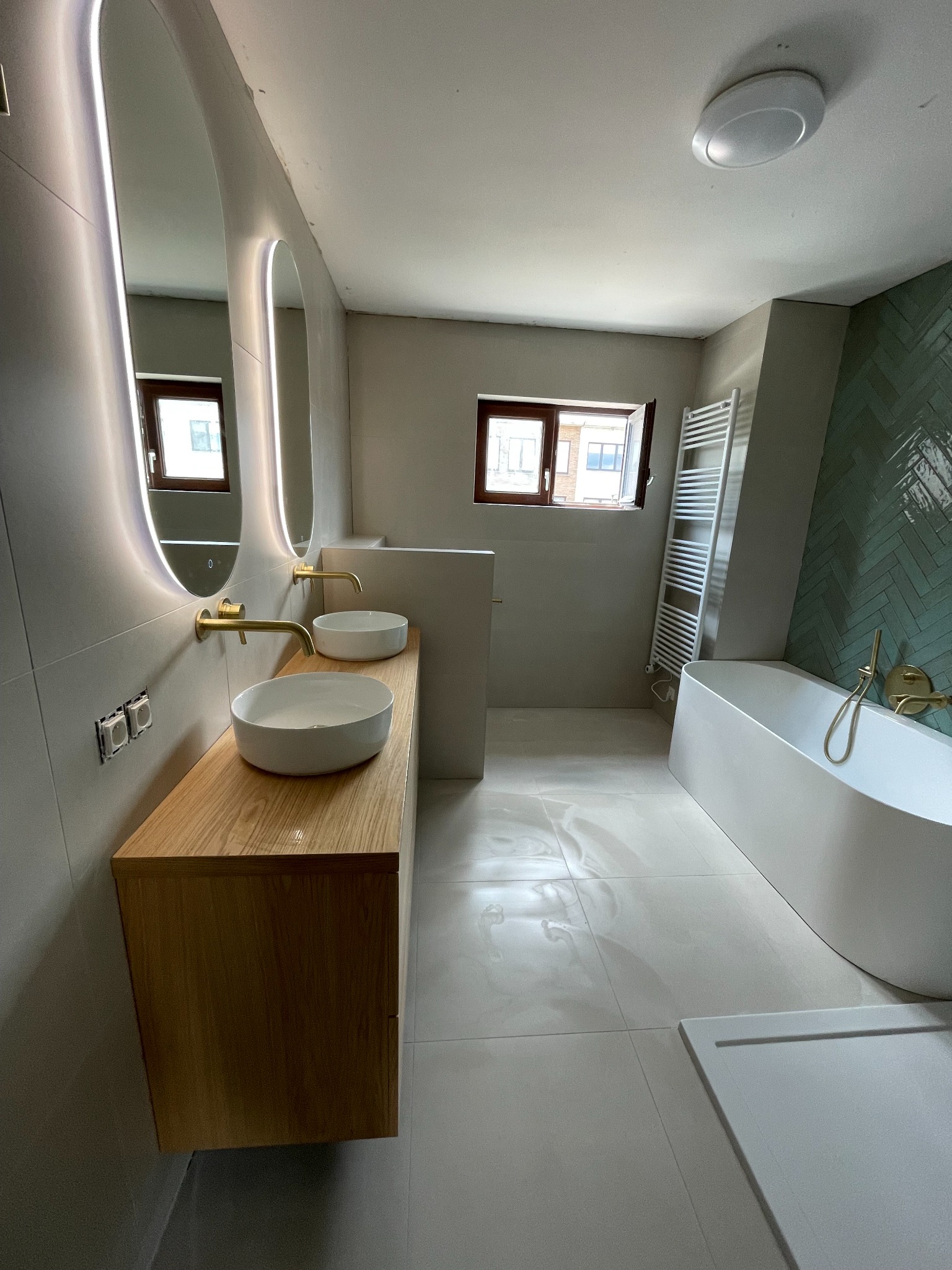 Nowoczesna łazienka z owalnymi lustrami podświetlanymi LED, drewnianą szafką pod umywalkami i zieloną ścianą z płytek ułożonych w jodełkę. Minimalistyczny design.