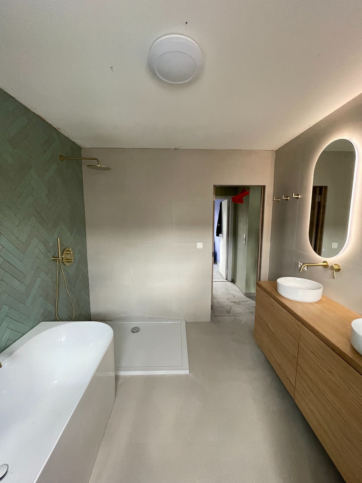 Nowoczesna łazienka z zieloną ścianą w jodełkę, złotą armaturą i podświetlanym lustrem. Widoczna wanna, kabina prysznicowa i szafka z umywalkami.