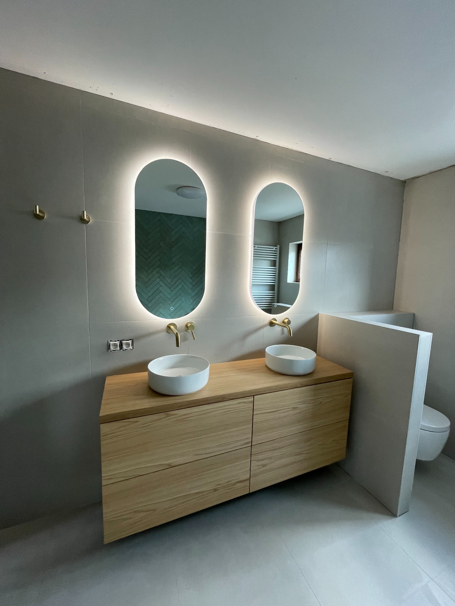 Nowoczesna łazienka z dwoma umywalkami nablatowymi, podświetlanymi lustrami owalnymi i drewnianą szafką. Minimalistyczny design, szare płytki i złote akcenty.
