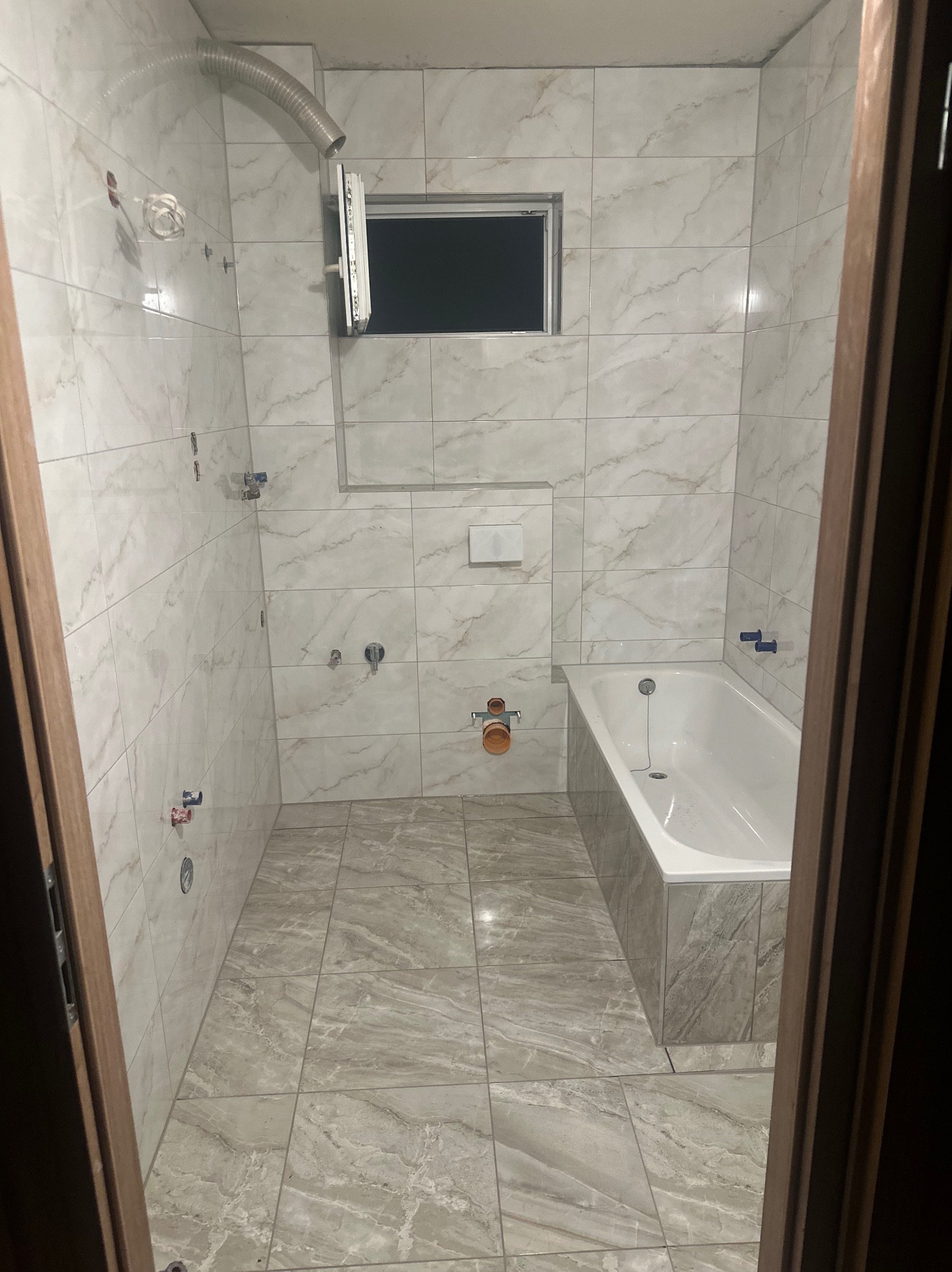 Wykończona łazienka z marmurowymi płytkami, białą wanną i oknem. Widoczne instalacje wodne i wentylacyjne. Ujęcie zza framugi drzwi.