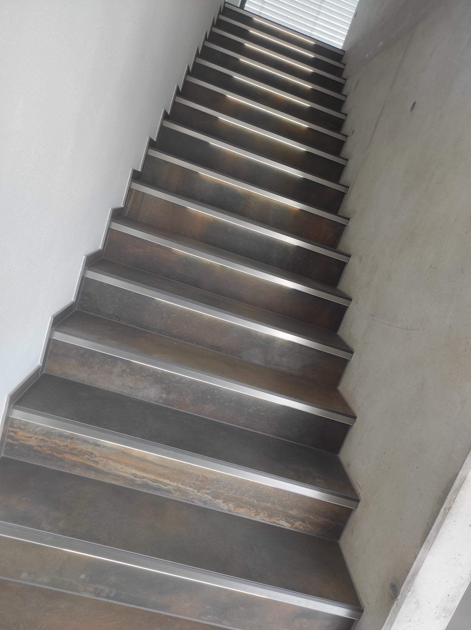 Stylowe schody z metalowymi krawędziami, prowadzące w górę, oświetlone naturalnym światłem z okna. Minimalistyczny design z surowym betonem w tle.