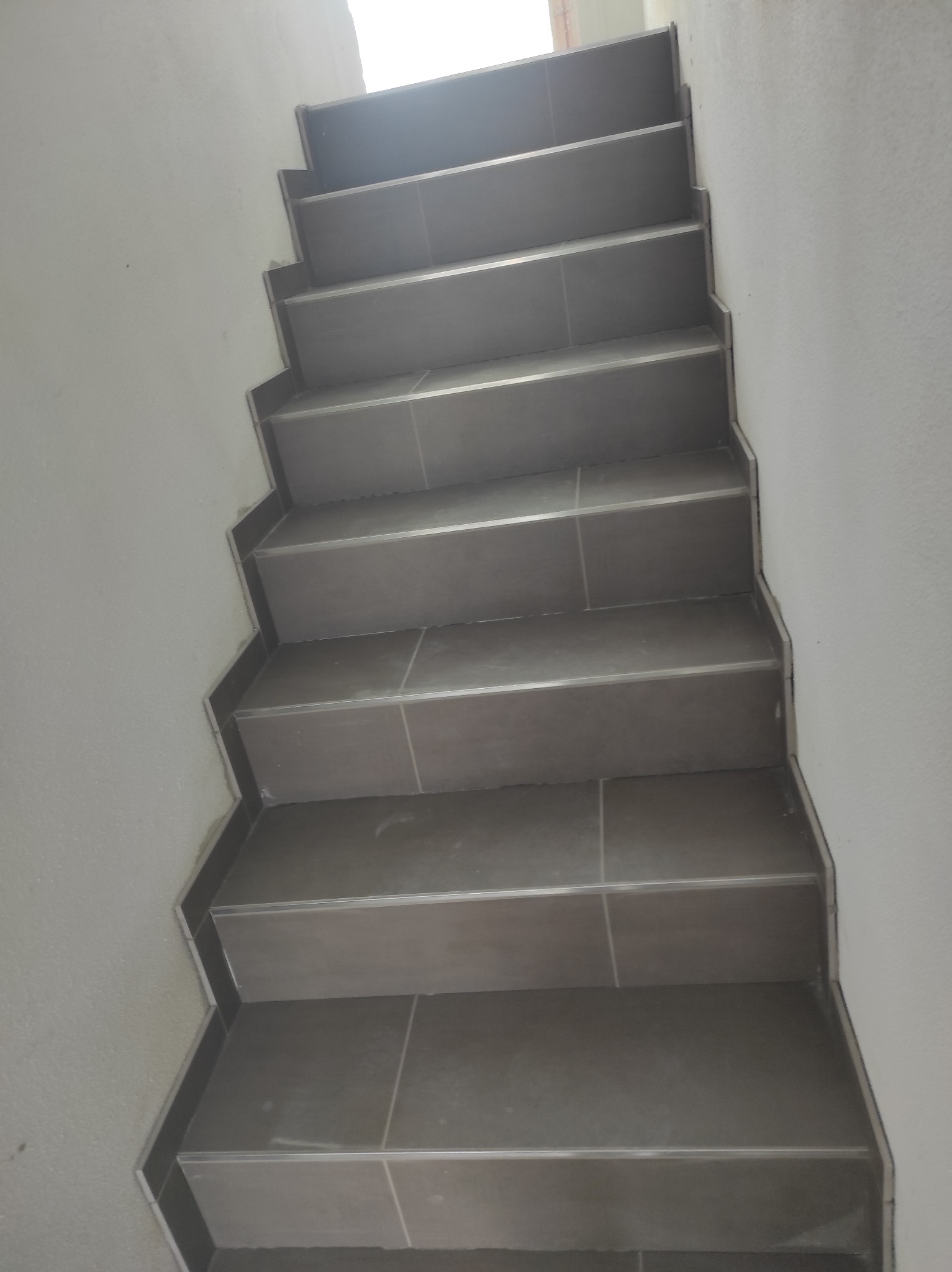 Schody wyłożone szarymi płytkami z metalowymi listwami, widok z góry, oświetlenie górne, ściany pomalowane na biało. Układanie płytek na schodach.