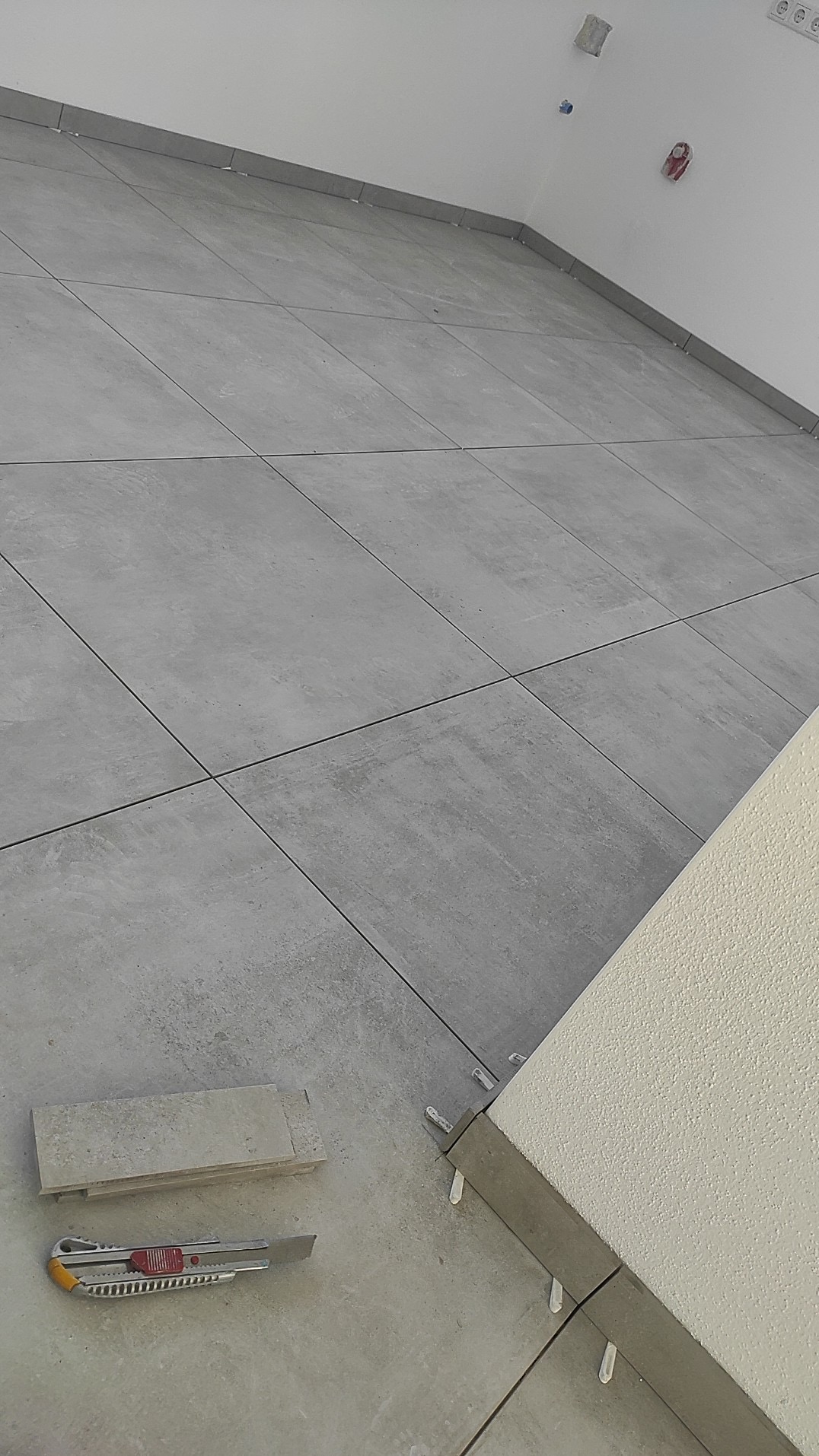 Ułożone szare płytki podłogowe z krzyżykami dystansowymi, nóż do cięcia płytek i fragment ściany z fakturą. Ujęcie z dołu, ukośne.