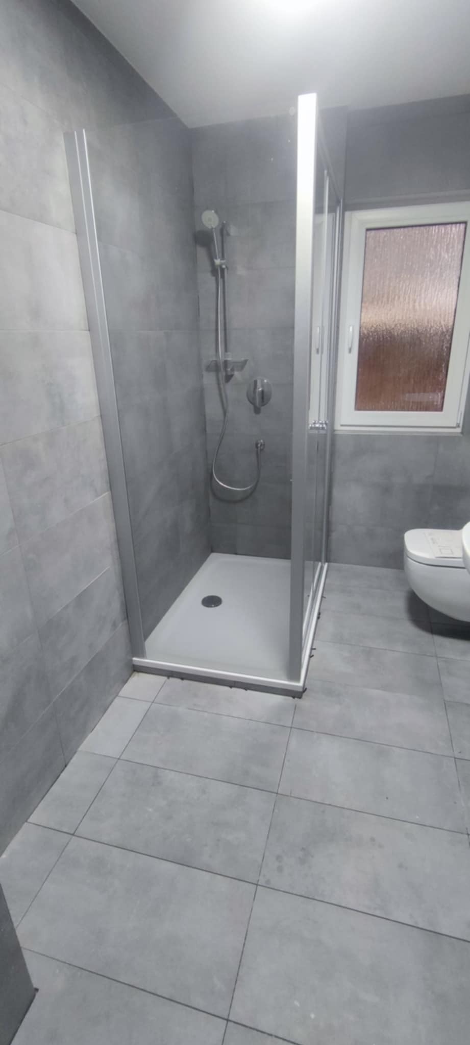 Szara łazienka po remoncie: kabina prysznicowa z brodzikiem, szare płytki na ścianach i podłodze, widoczna armatura i toaleta. Minimalistyczny design.