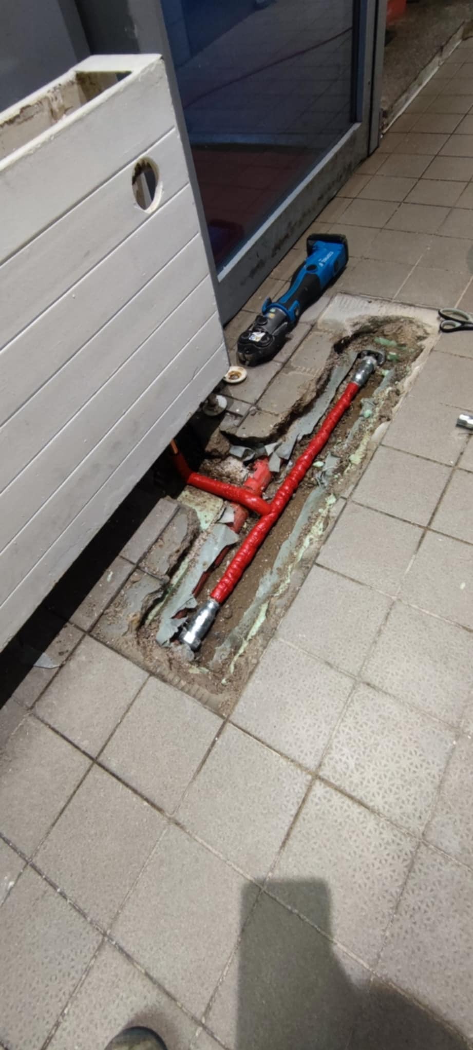 Czerwone rury instalacji hydraulicznej w wykopanym otworze w podłodze z płytek, obok narzędzie do zaciskania rur i biała osłona.