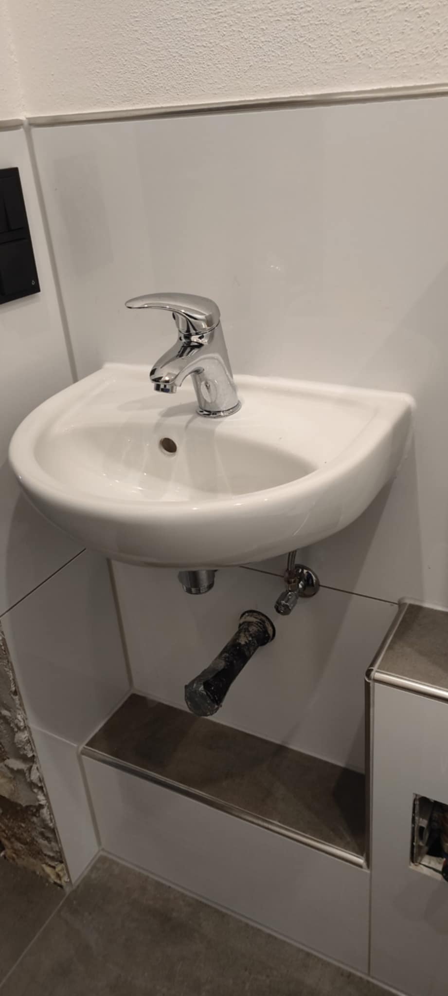 Biała umywalka z chromowaną baterią, widoczne rury odpływowe pod spodem. Fragment ściany z płytkami i niedokończone wykończenie w tle.