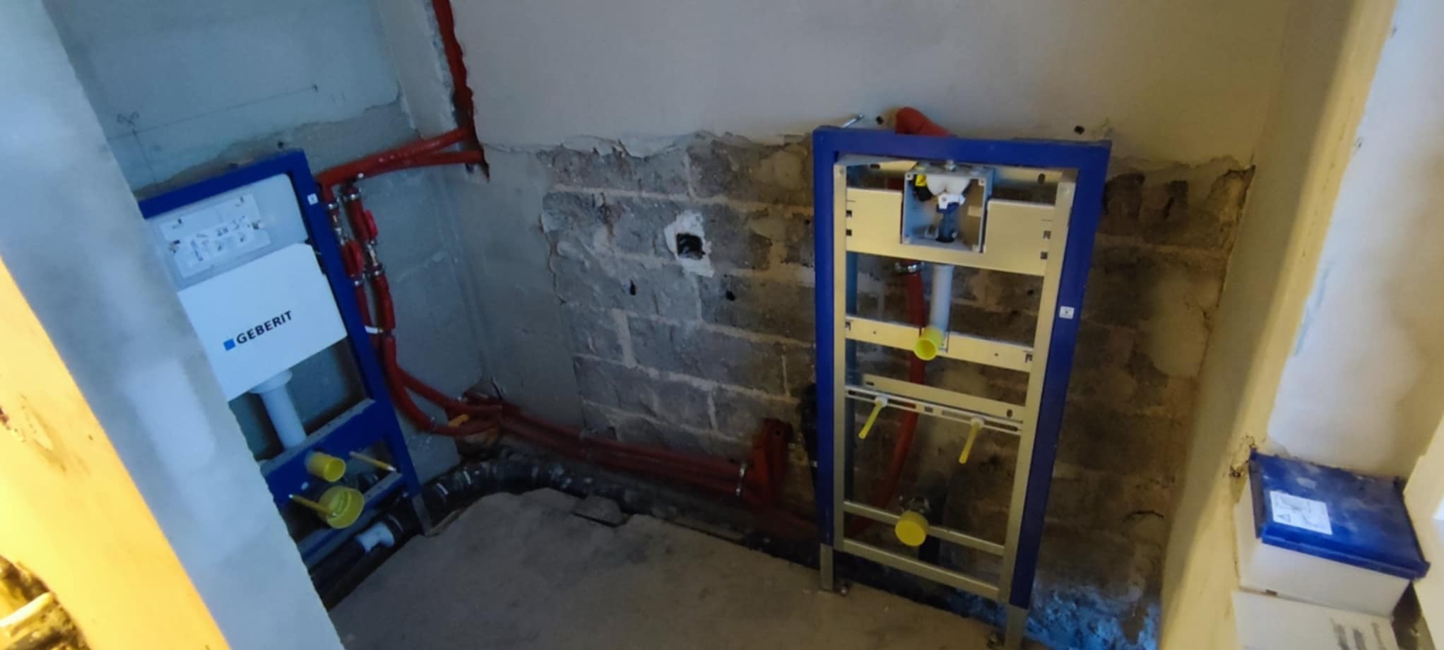Dwa stelaże podtynkowe Geberit z czerwoną instalacją hydrauliczną na tle surowych ścian w trakcie remontu łazienki. Widoczne elementy mocujące i rury.