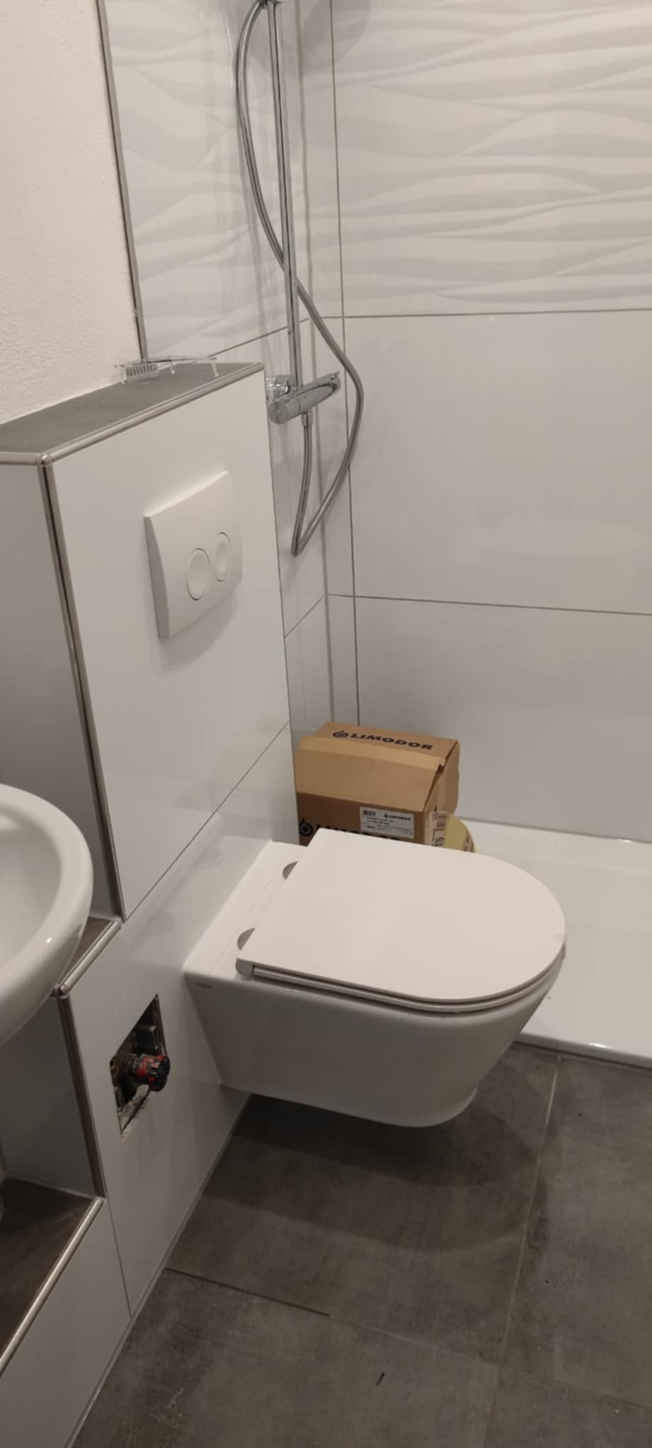 Biała, podwieszana toaleta z zabudowaną spłuczką i przyciskiem, obok umywalka. Widoczna instalacja wodna i karton Limodor. Nowoczesna łazienka po montażu.