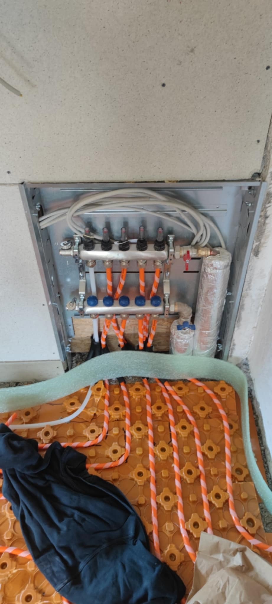 Rozdzielacz ogrzewania podłogowego z rurami w izolacji, zamontowany w metalowej skrzynce, widok z góry, częściowo zakryty czarnym materiałem.