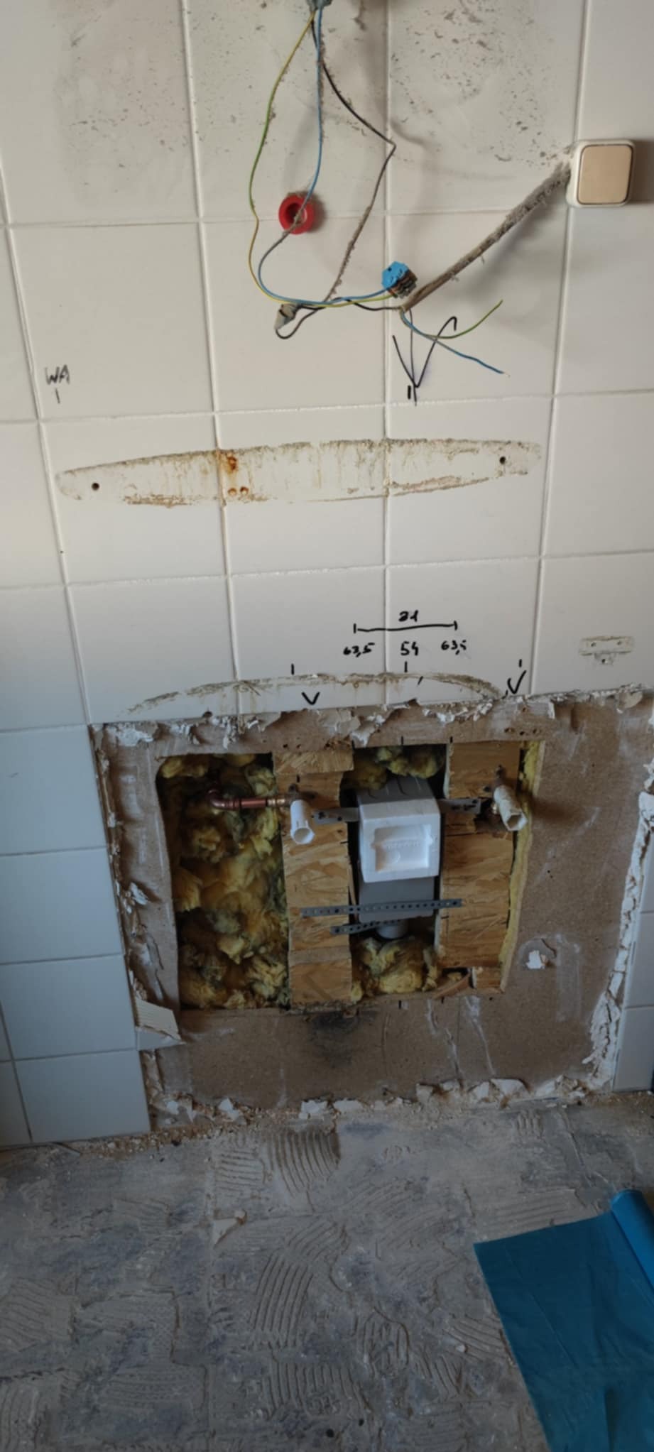 Ściana z odsłoniętą instalacją hydrauliczną w trakcie remontu łazienki. Widoczne rury, izolacja i fragmenty płytek. Na ścianie odręczne notatki z wymiarami.