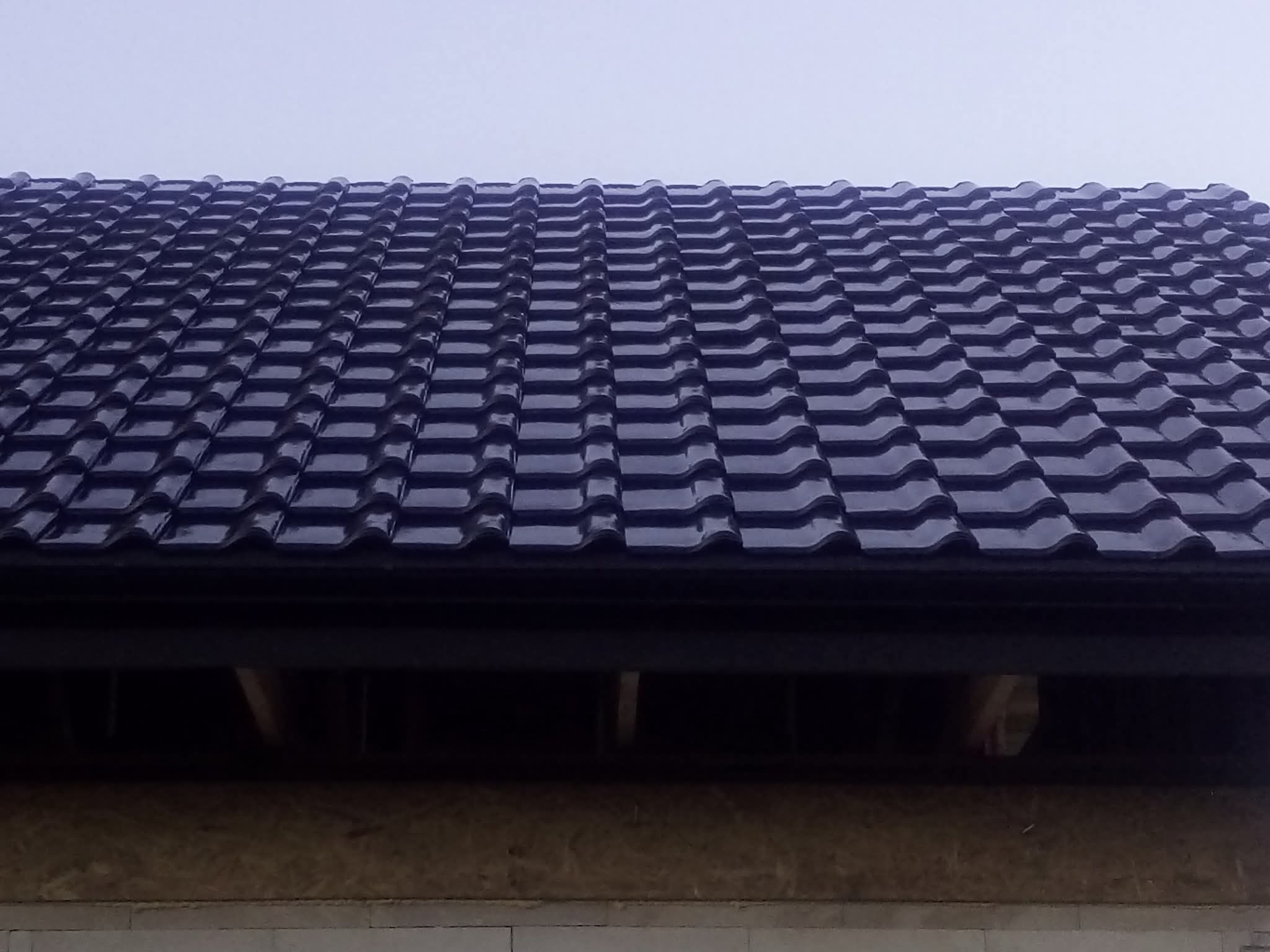 Błyszczący, ciemny dach z dachówki ceramicznej, widoczny z dołu, z fragmentem rynny i ocieplonego poddasza. Ujęcie z bliska, pochmurne niebo w tle.