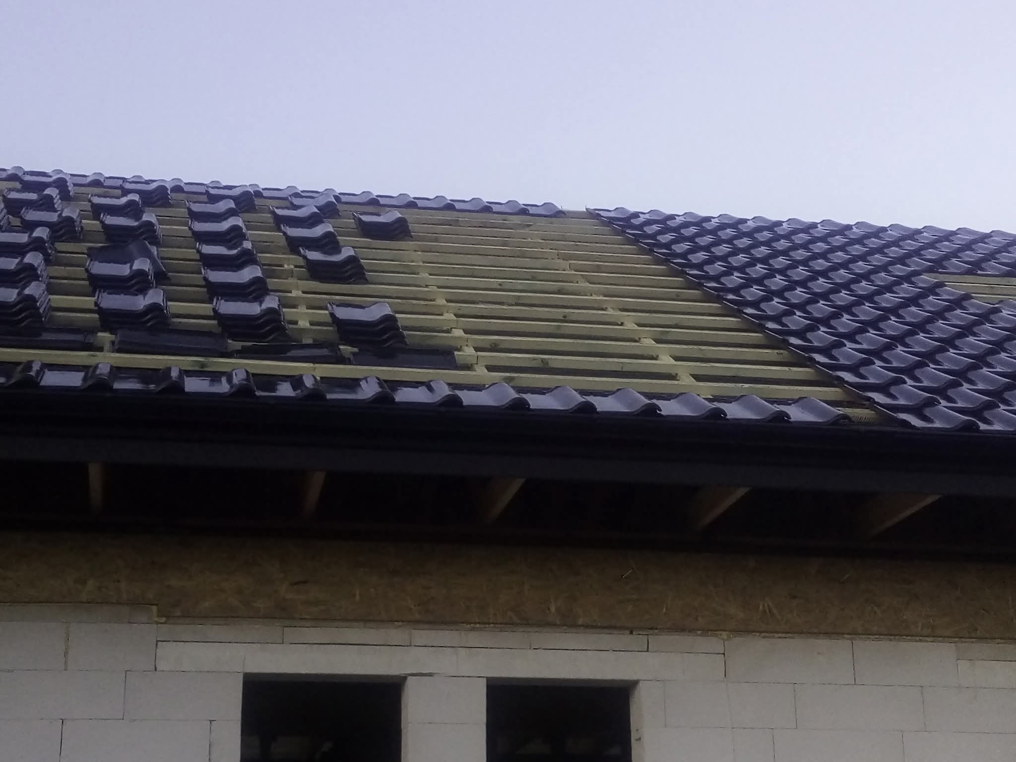 Układanie ciemnej dachówki na drewnianej konstrukcji dachu, częściowo pokrytego. Widoczny fragment ściany budynku z pustaków.