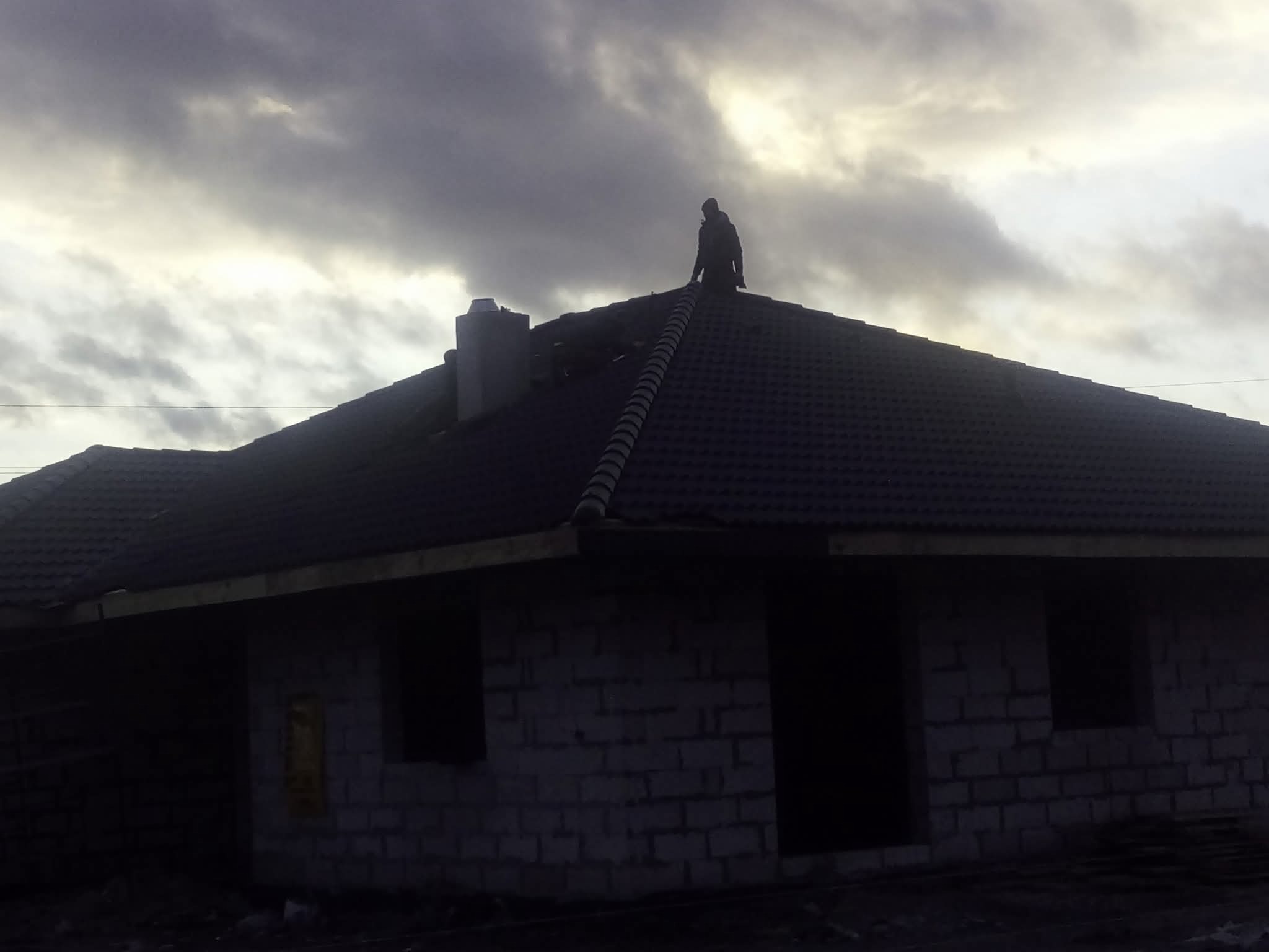 Dekarz na dachu domu w trakcie budowy, widoczny komin i fragment ściany z pustaków, pochmurne niebo w tle. Praca na wysokości.