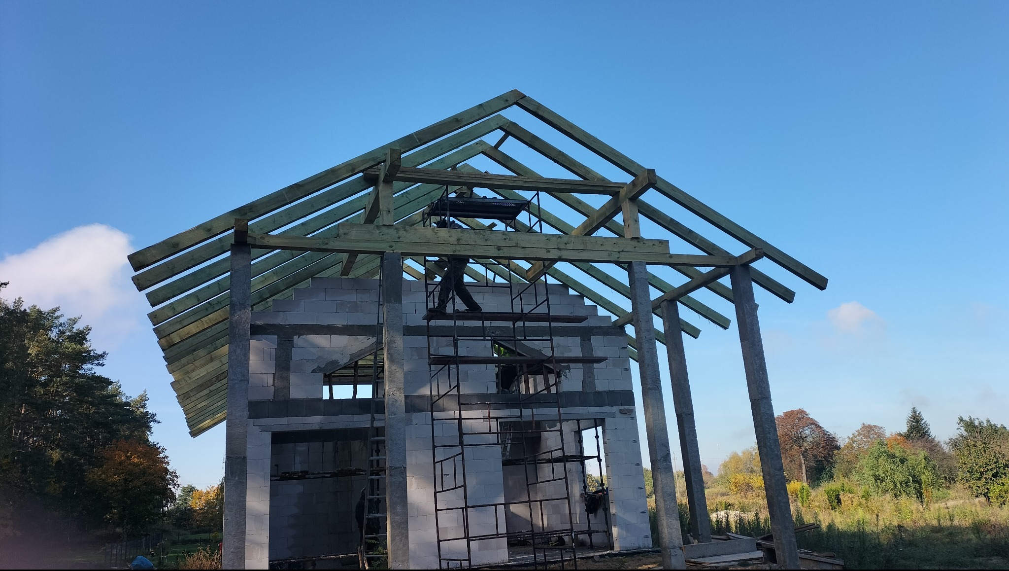 Wznoszenie więźby dachowej na budynku z betonu komórkowego, widok z dołu na konstrukcję i pracownika na rusztowaniu, błękitne niebo w tle.