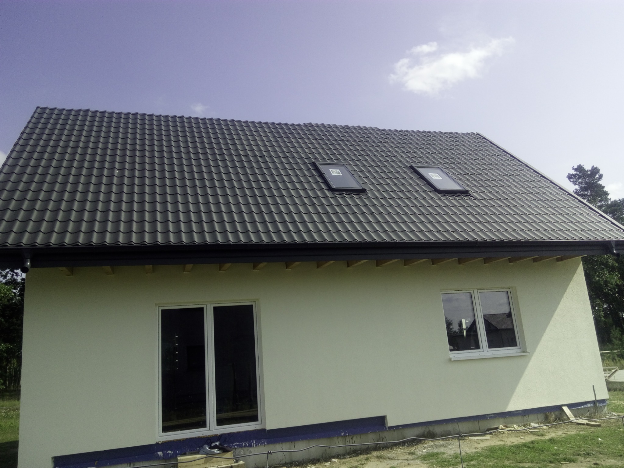 Nowy, grafitowy dach z dwoma oknami połaciowymi na jasnym domu jednorodzinnym w Rzęczkowie. Widoczne rynny i fragment elewacji.